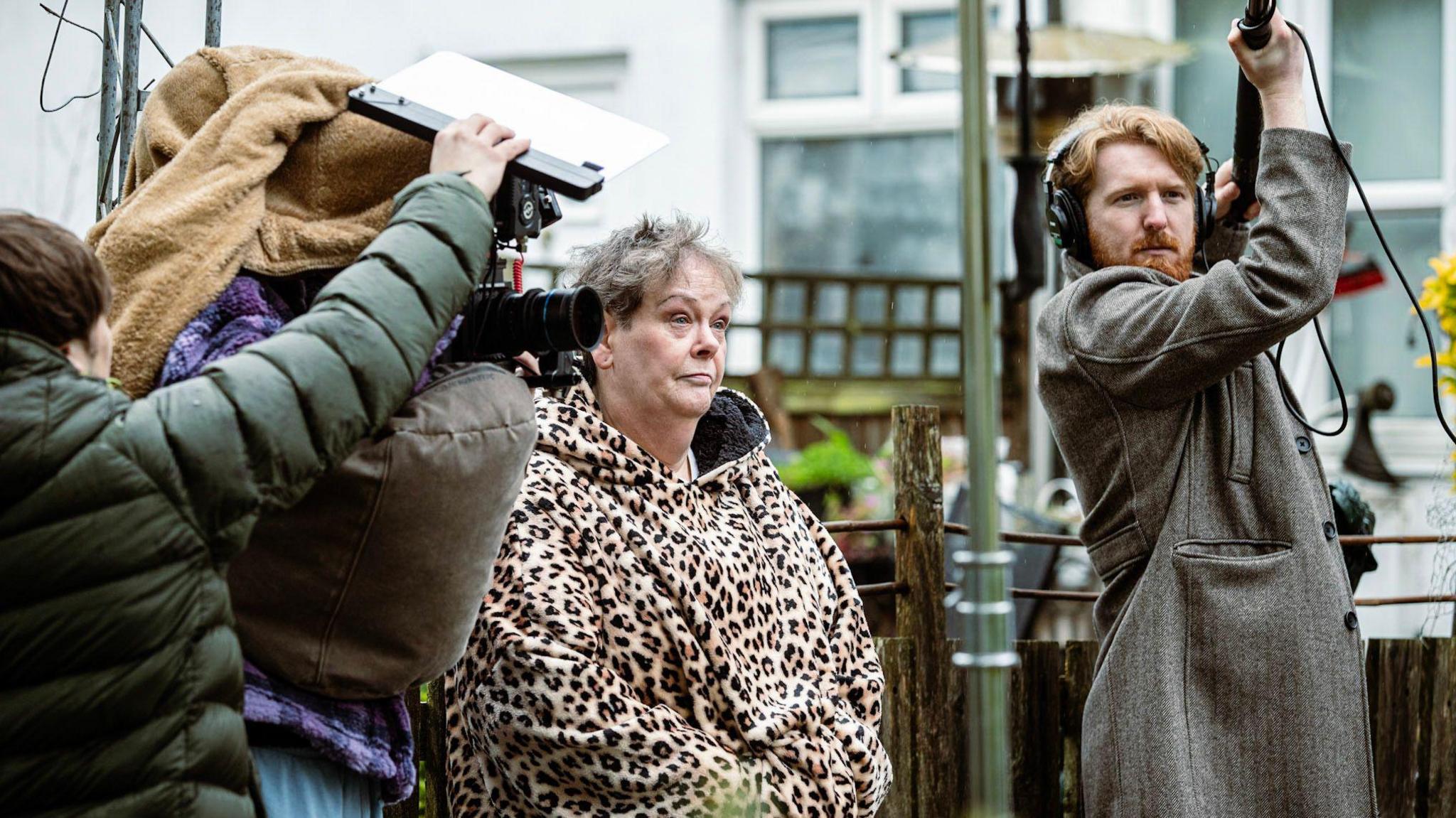 A behind-the-scenes image of Anne Hegerty. She is wearing a leopard print hooded top. She is standing in a back garden. A man with a boom microphone is stood near her and two people are operating a film camera.