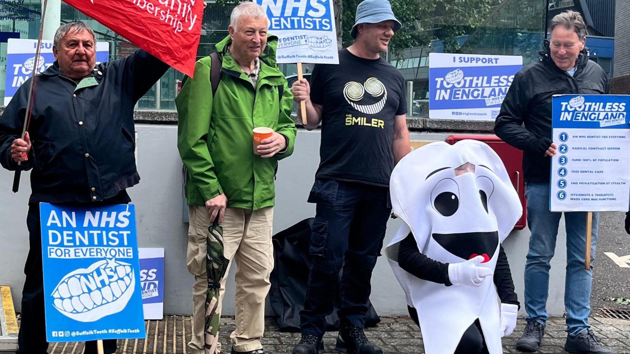 Six member of the Toothless in Suffolk campaign group stand outside Endeavour House. They are equipped with posters and flags.