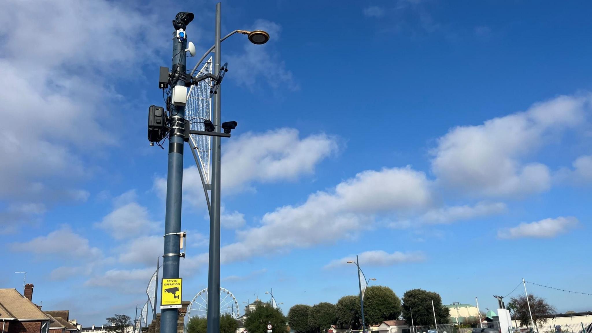 Tall pole with camera and sound devices attached on a street in Great Yarmouth with the Ferris Wheel in the background.