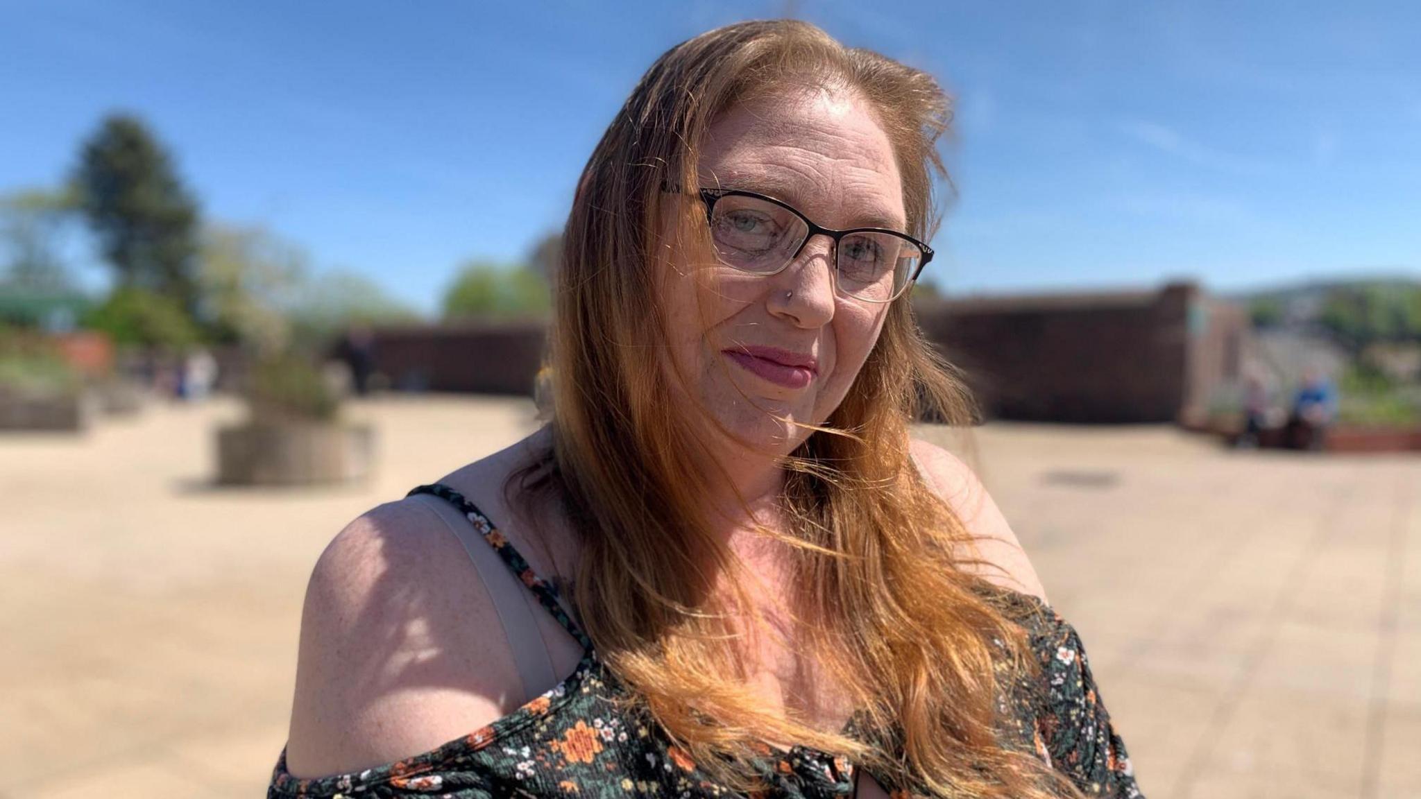 Melanie Harris in black summer dress with shoulder straps and floral print. She wears black-rimmed glasses and has long red hear worn loose. She is pictured outside in sunny weather