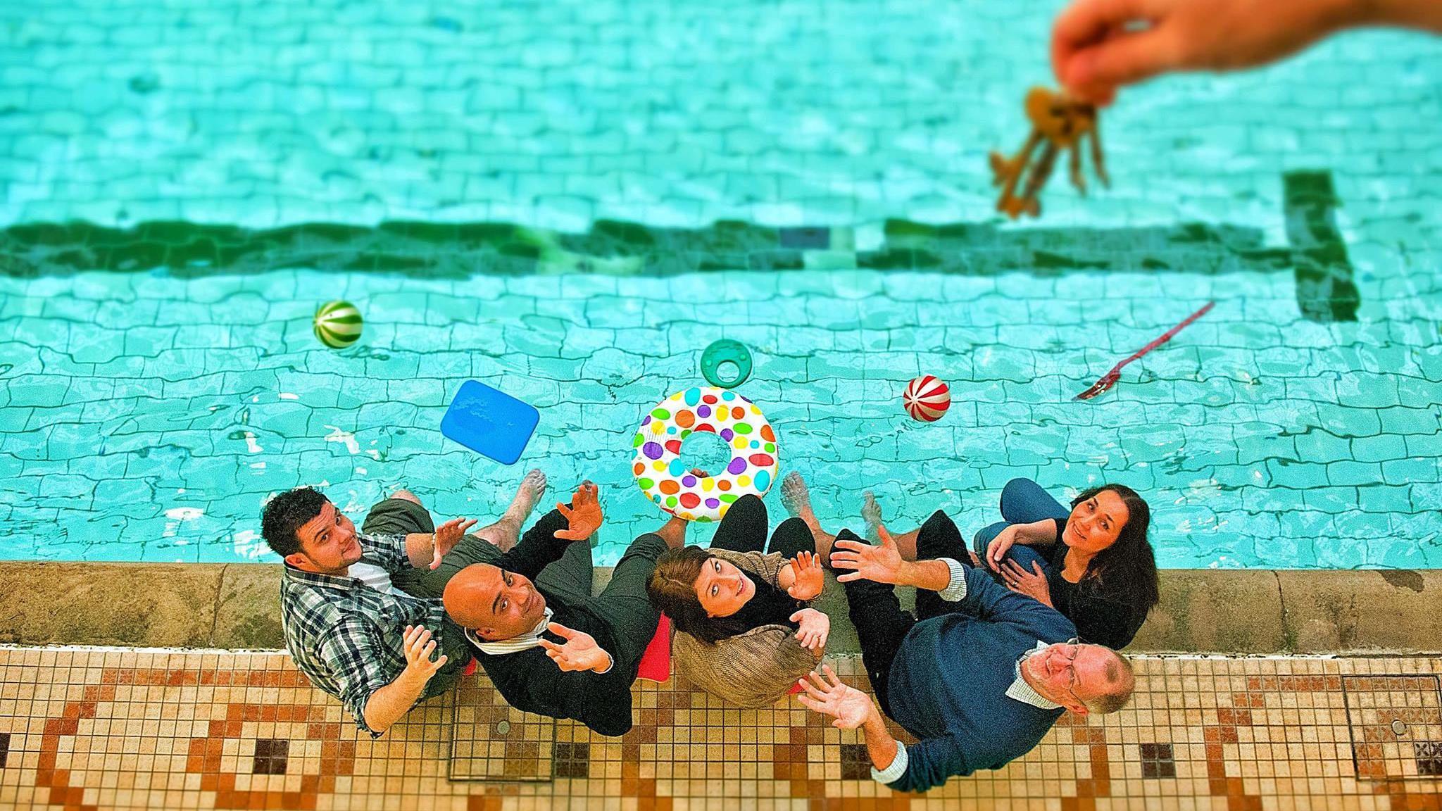 A group of people sit on the side of a swimming pool and look up towards hanging keys