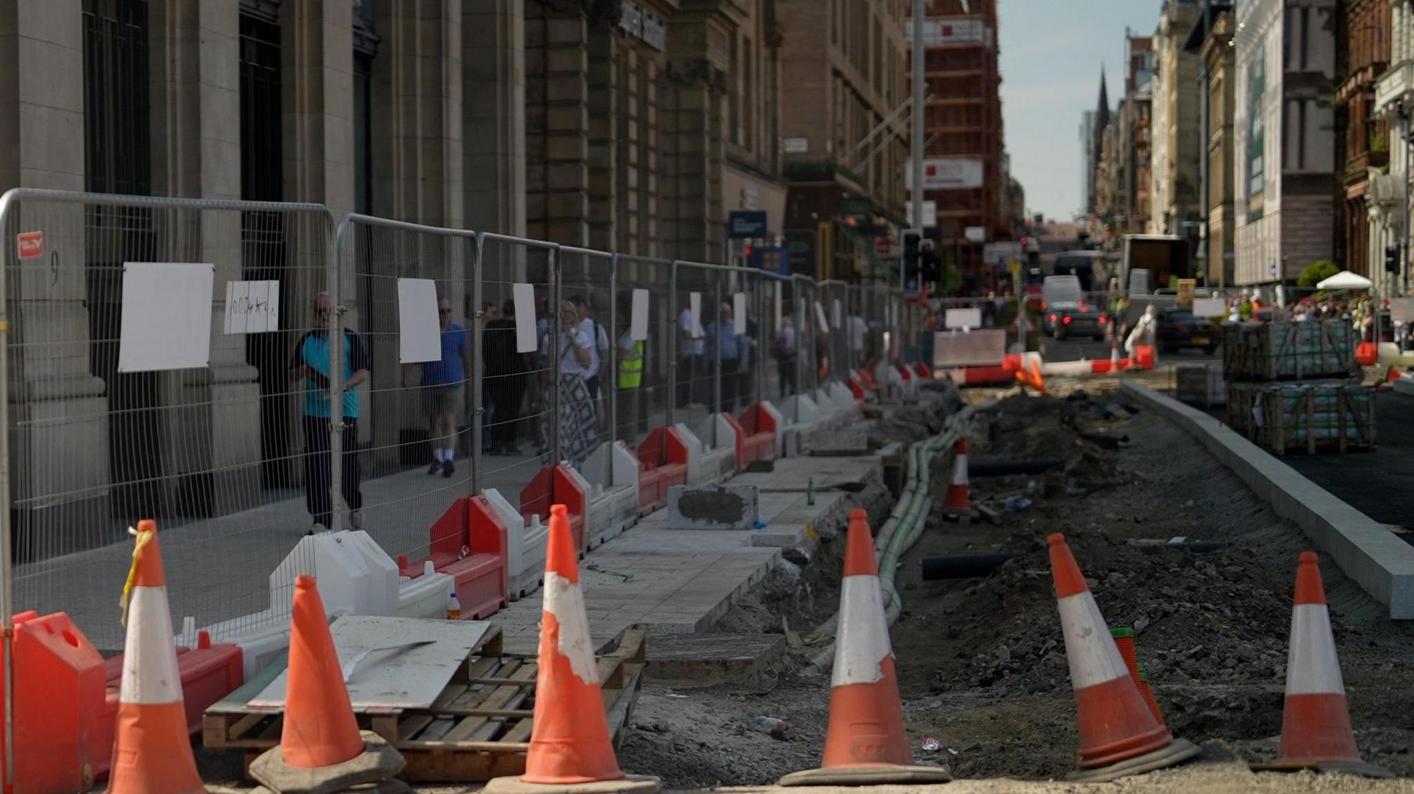 Roadworks along a city centre street with traffic cones, safety barriers and fenced-off excavations. Pedestrians walk along a temporary pavement beside construction works, with buildings and traffic visible in the background.