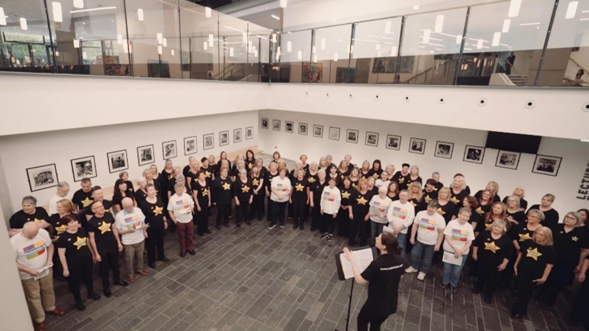 Members of the Dementia and Rock Choir in black and white t-shirts performing The Climb being led by choir leader, Rachel Coulter. 