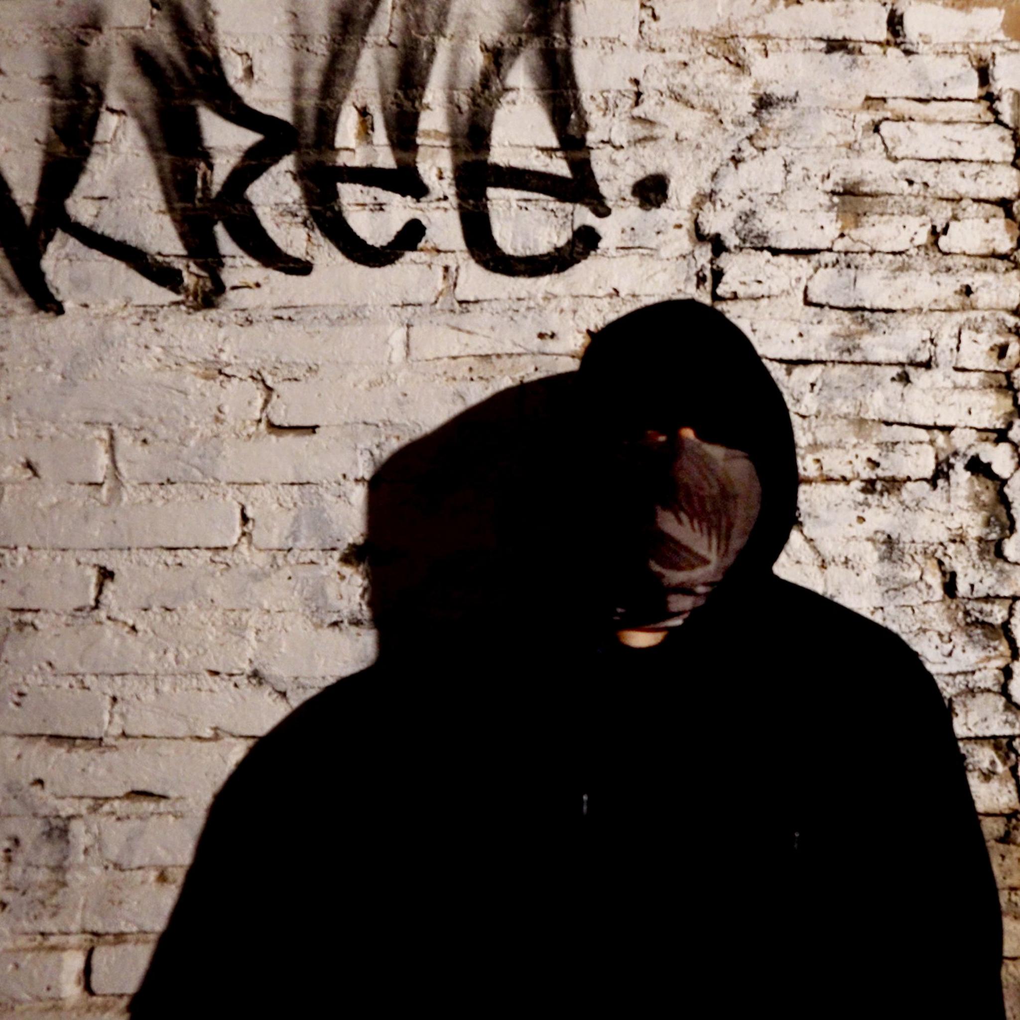 A man standing in shadow and further concealed by a black hoodie and a bandana over his face, in front of the wall of a cartel safe house, which has crumbling brick and fading white paint. There is unintelligible graffiti above his head.