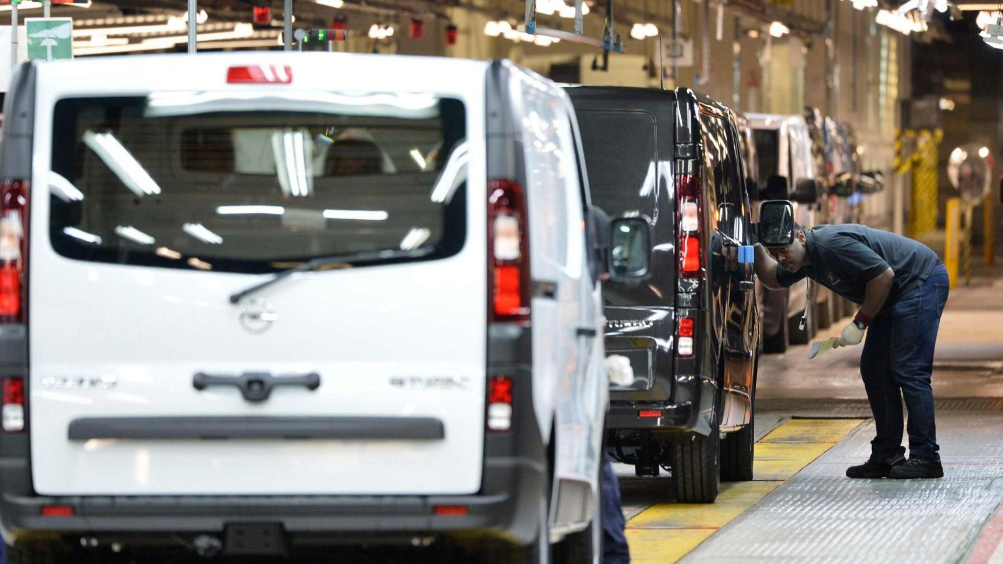 A Vauxhall work inspects a black van on the production line in Luton. The rear of a white van is in the foreground.