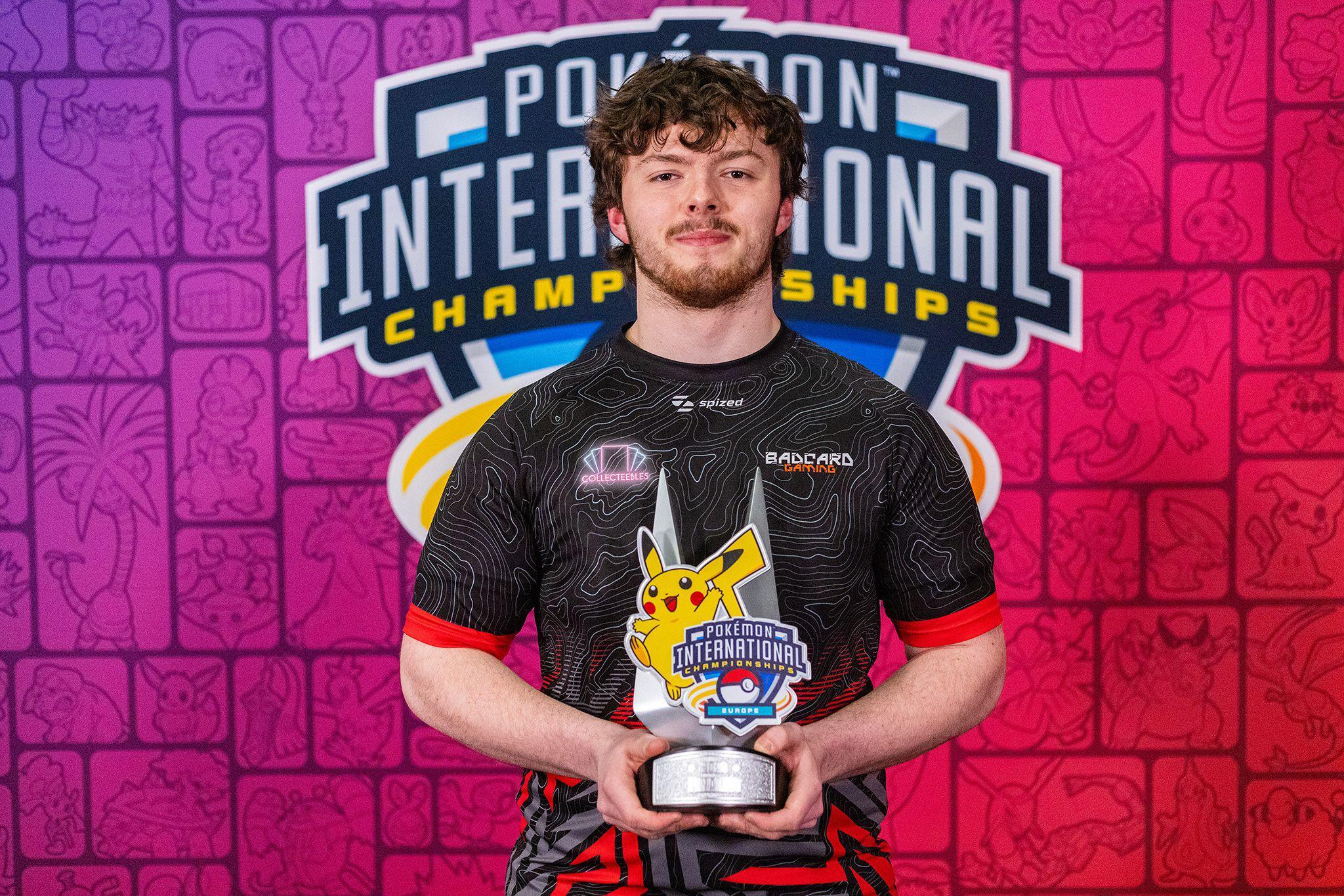 Drew Stephenson stands in front of a "Pokémon International Championships" logo on a pink-coloured wall. He's wearing a black and red team shirt and cupping a championships trophy - featuring the tournament logo and a 2D image of Pikachu - in his hands at waist level.