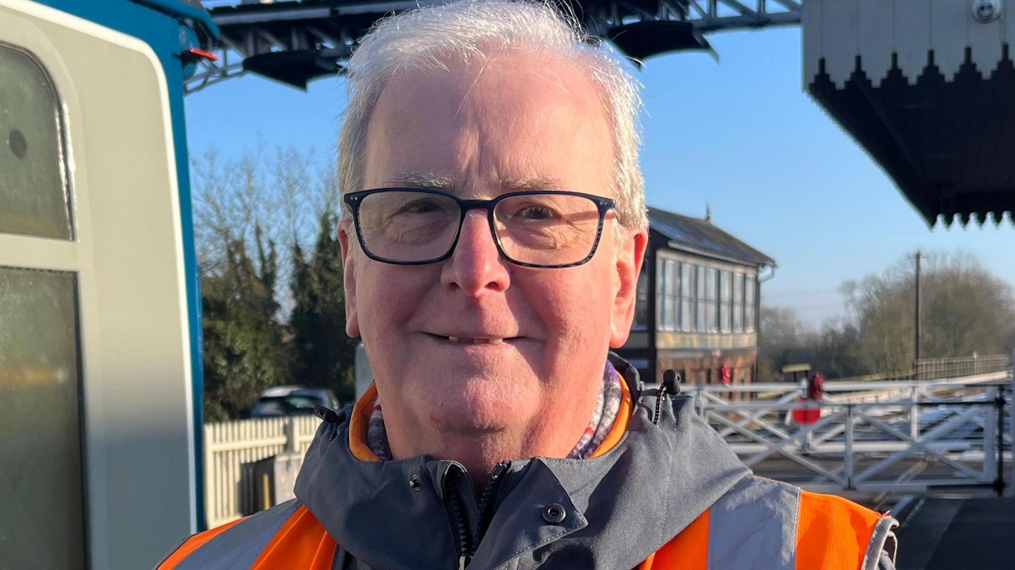 Michael Purcell is standing to the side of a train and smiling at the camera. He has grey hair, square framed glasses and is wearing an orange high vis vest. It is blue sky behind him and the light from the sunrise is shining on his face. 