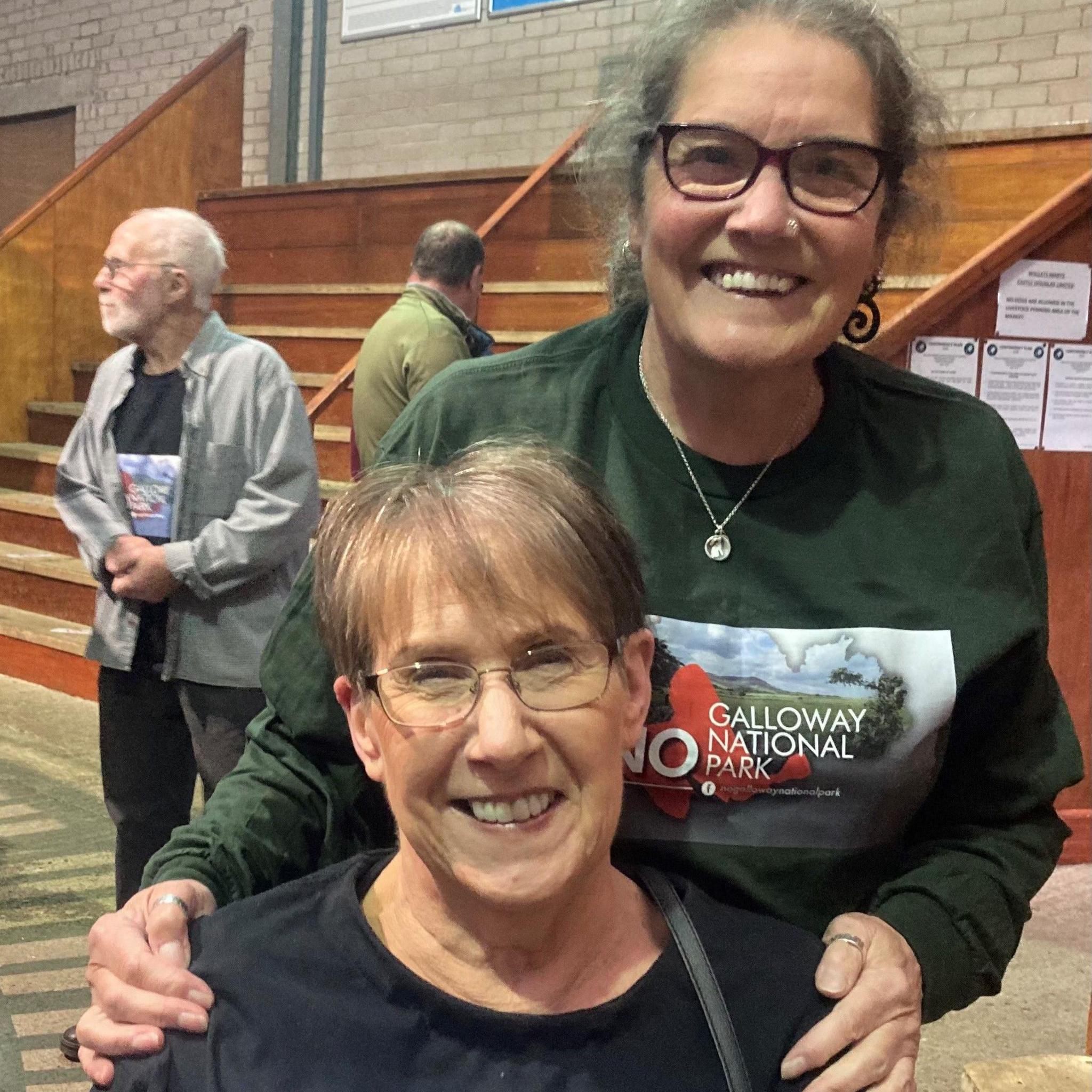 Two women - one seated and one standing - both wearing glasses and No to the Galloway National Park jumpers