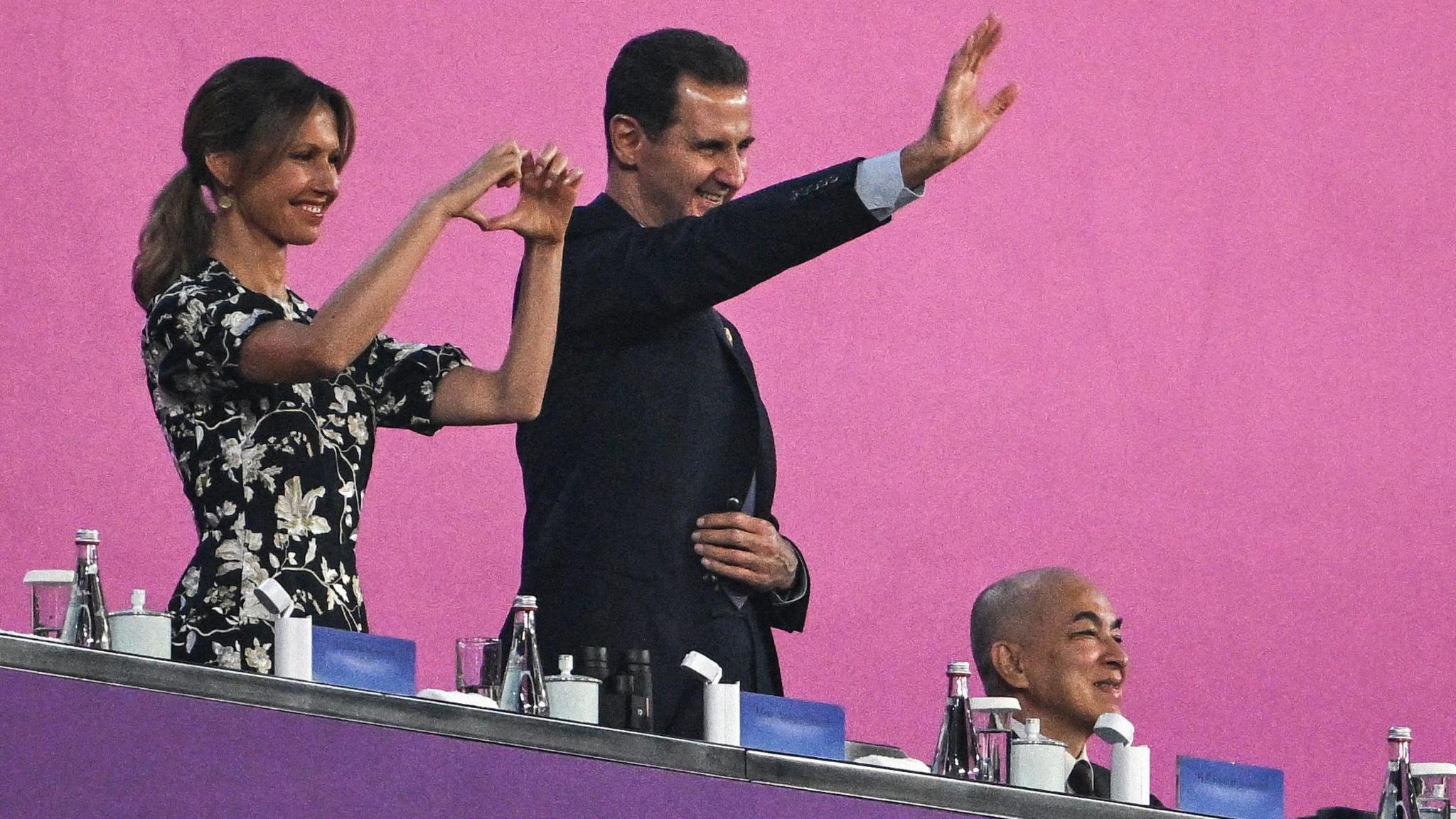 Asma al-Assad makes a heart with her hands and smiles, beside Bashar al-Assad waving and smiling, in front of a pink background