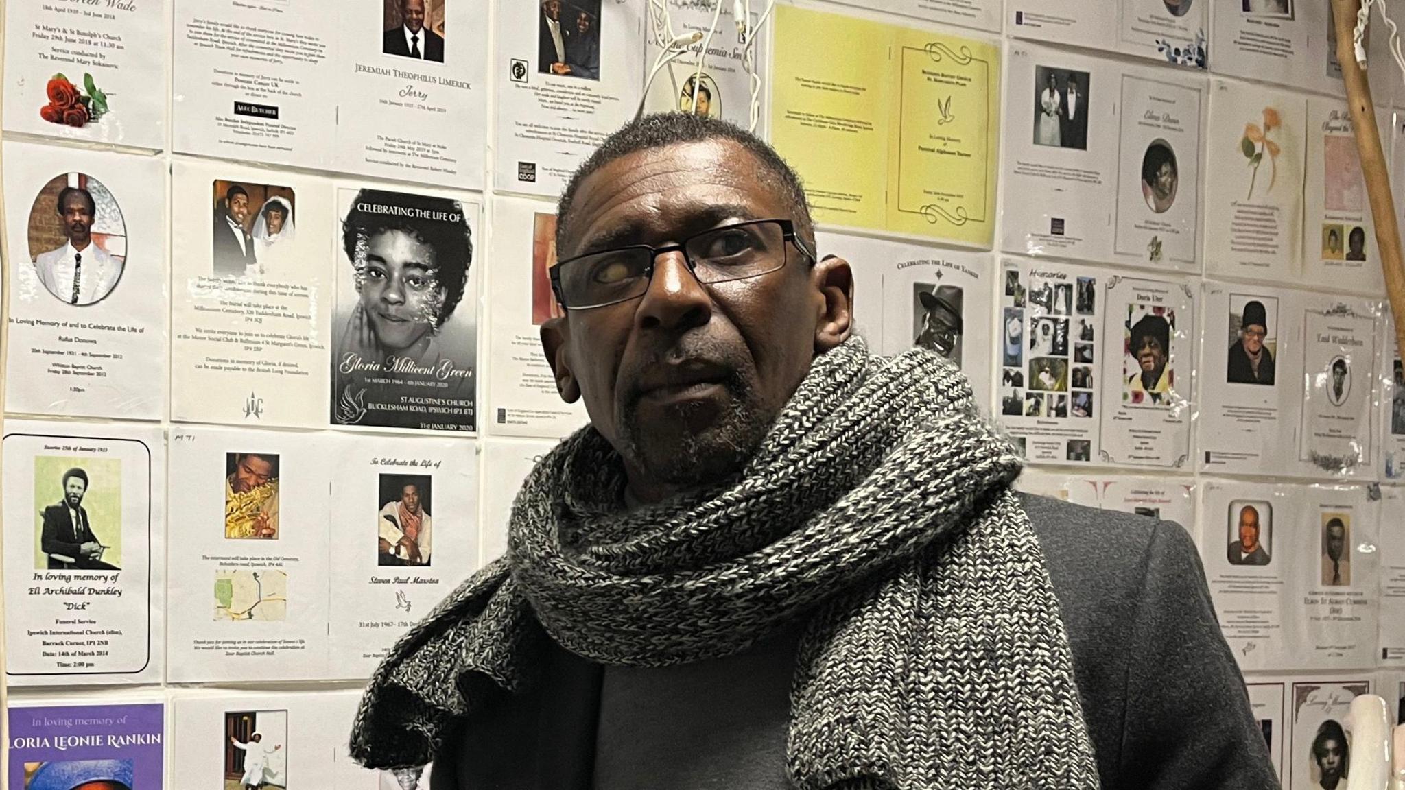 Max Thomas from Ipswich Windrush Society looks at the camera. He has a thick grey scarf around his neck and wears a dark suit jacket.