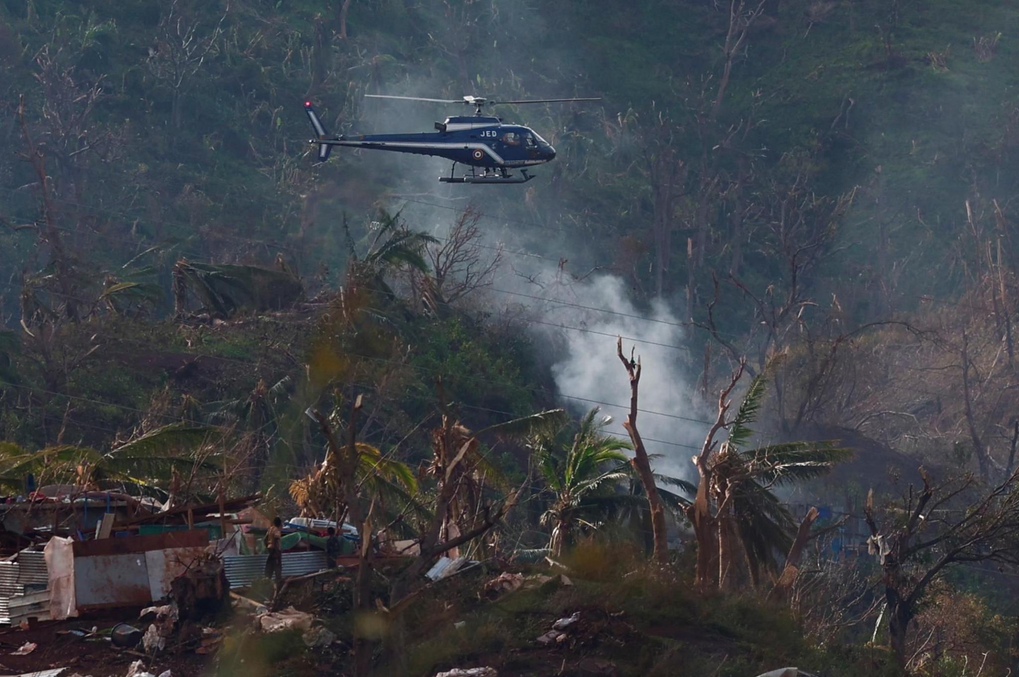 A helicopter from the French Gendarmerie flies over the Longoni village in the aftermath of Cyclone Chido in Mayotte. Trees and buildings are flattened or badly damaged and smoke rises from the ground, from where two people inspect the damage.
