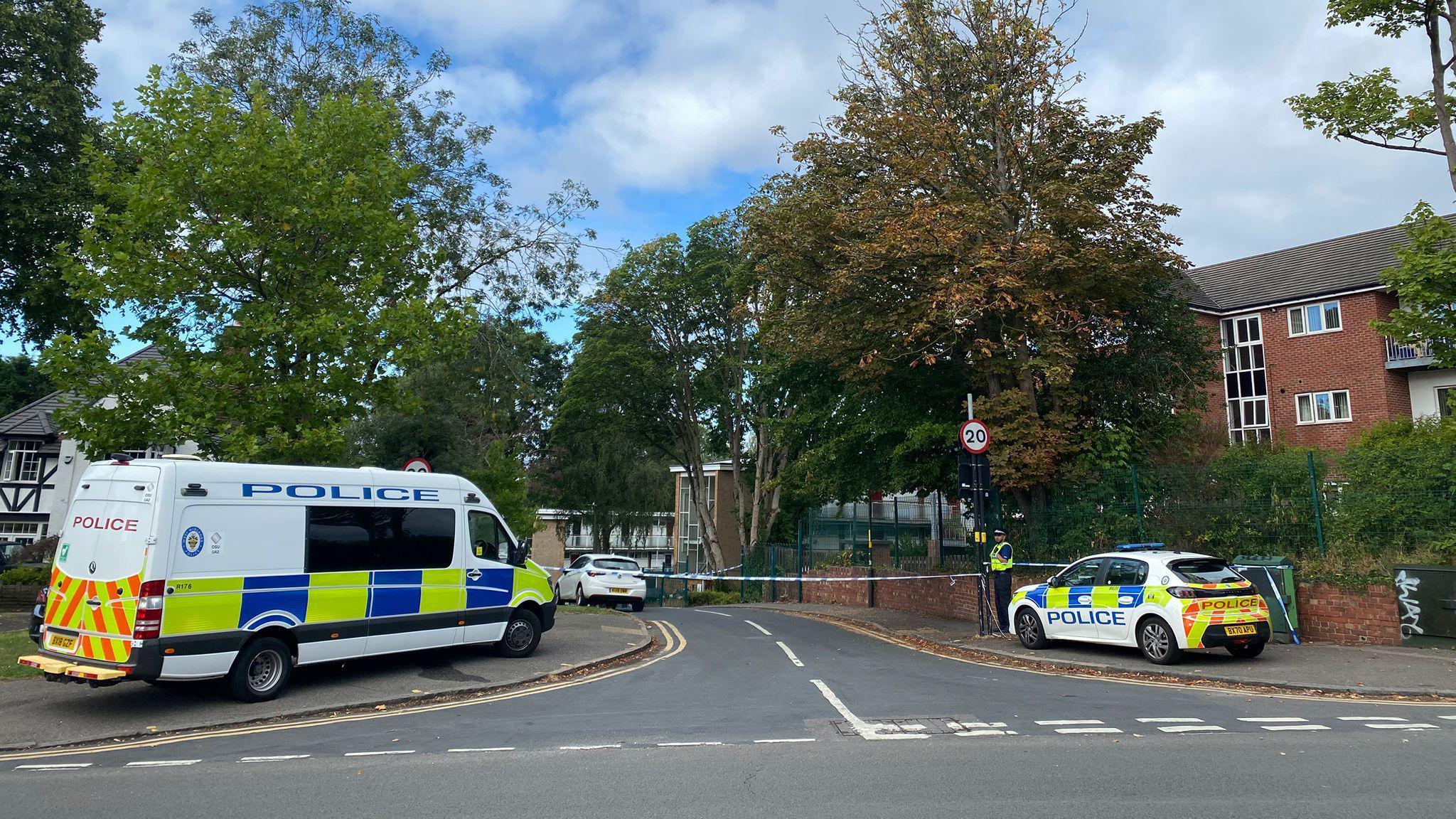 A road with blue and white police tape across it. There is a police van parked on the left and a police car on the right. A policeman is guarding the tape