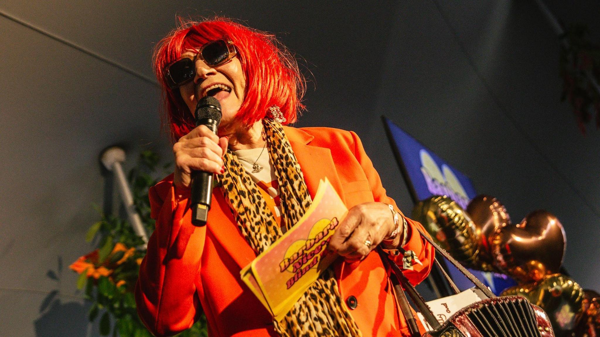 Mrs Barbara Nice wears an orange jacket, leopard-print scarf, dark glasses and a red wig