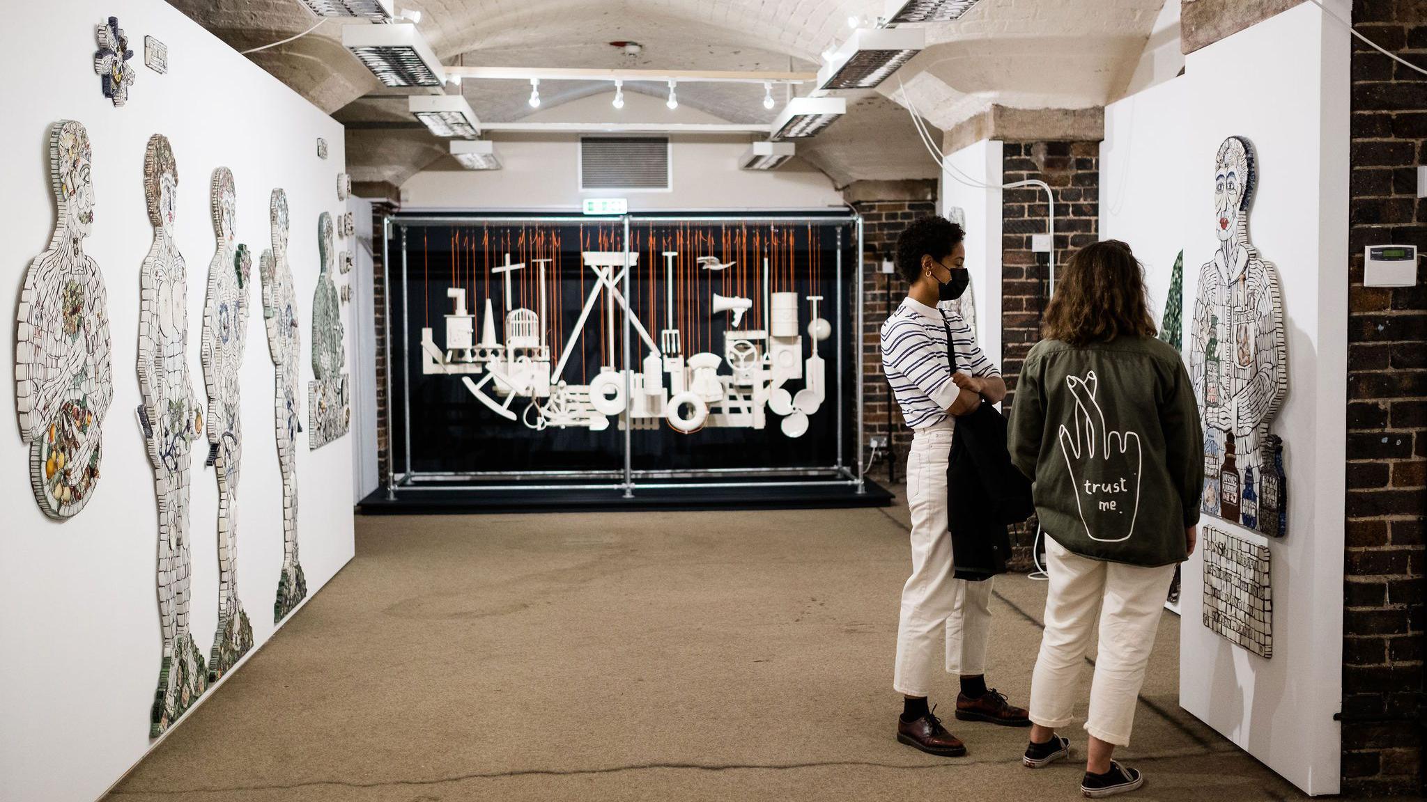 Two people look at an art display of a person mounted on a white wall. The room they're in features other displays with whit shapes in the distance and more artworks of people on the left.