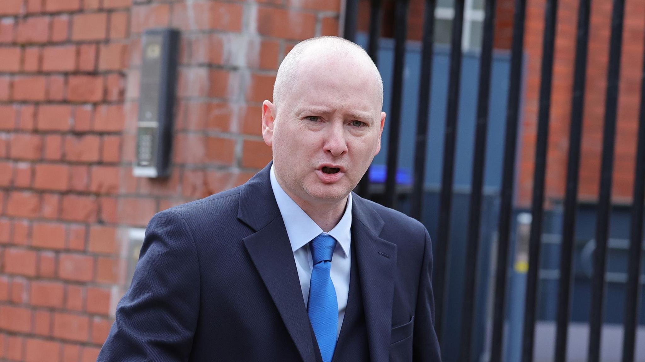A bald headed man in a dark suit with a blue shirt and tie