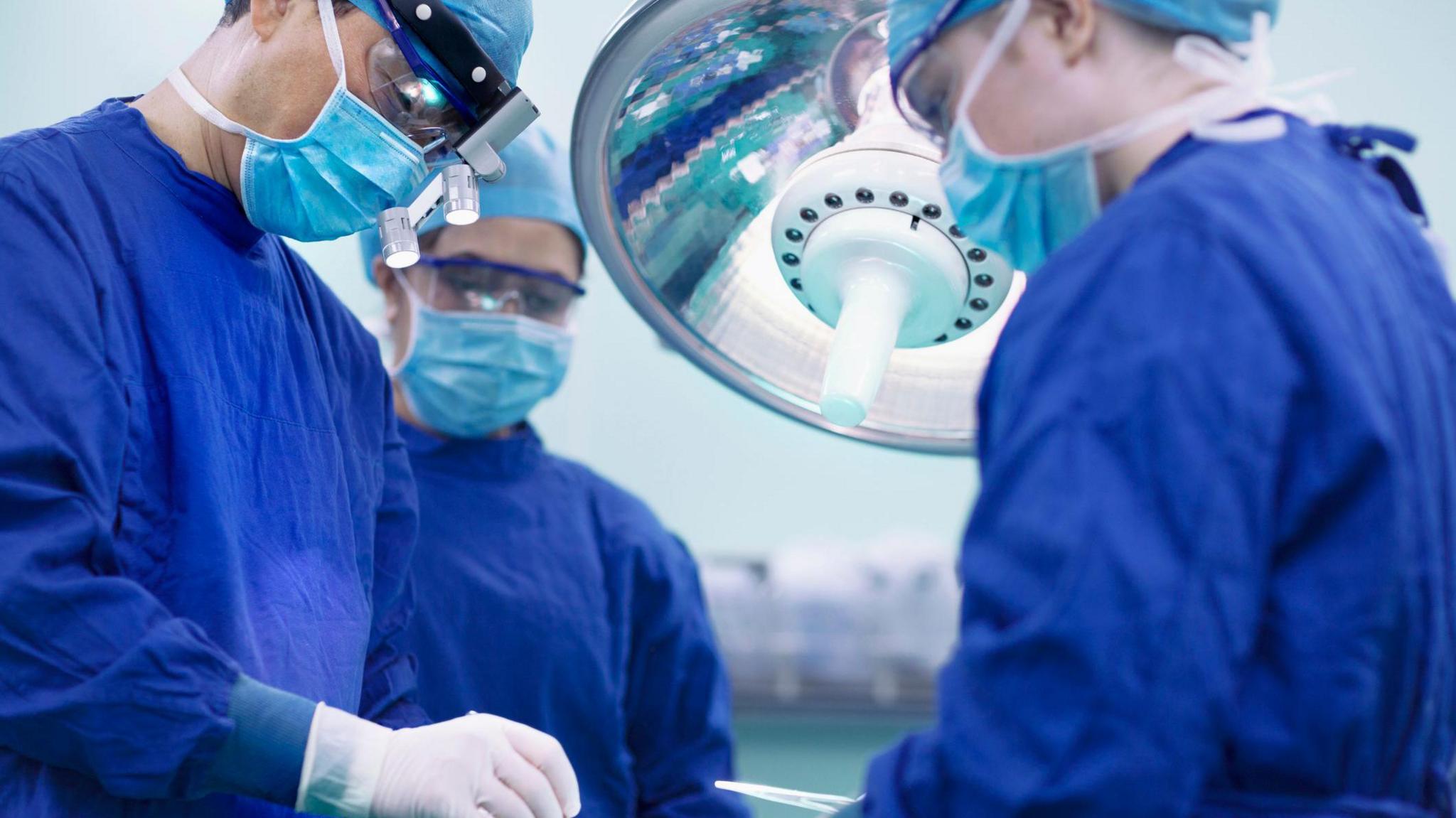 Surgeon with a head light on and blue mask performs a surgery out of sight of the camera. There are two other surgeons dress in blue robes beside him.