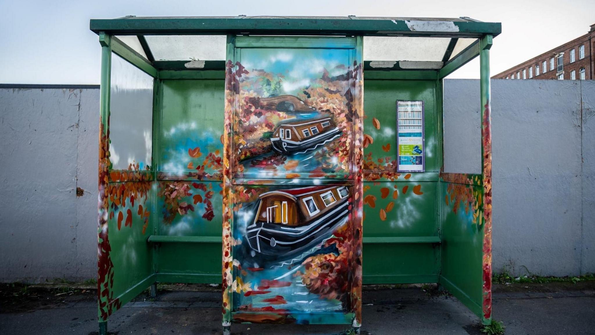 A green bus shelter with the central panel painted with two brown, blue and red canal boats. They are on water and surrounded by leaves.  