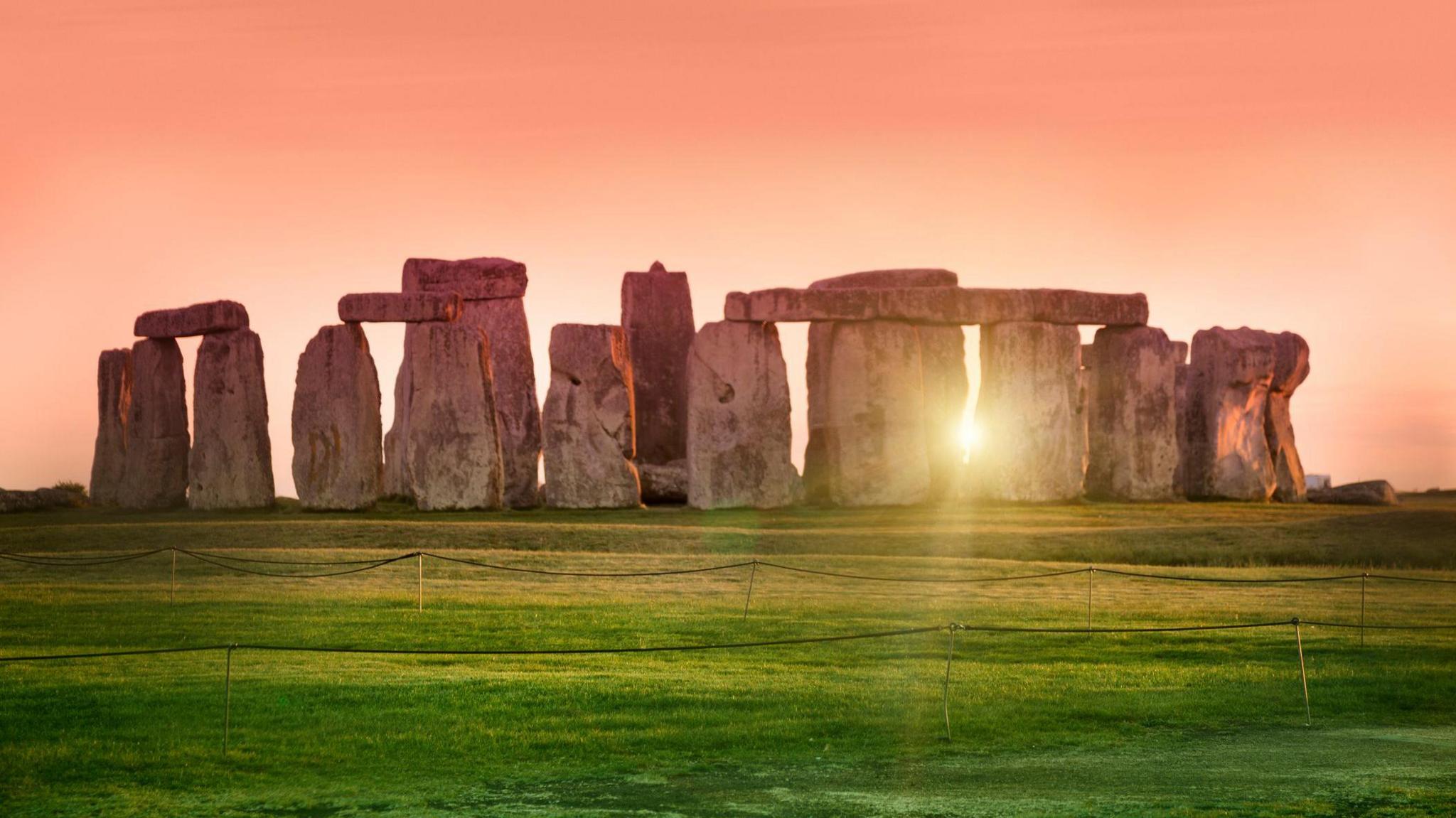 An image of the Stonehenge at sunrise. Sunlight is streaming through the stones, with green grass and pink skies visible