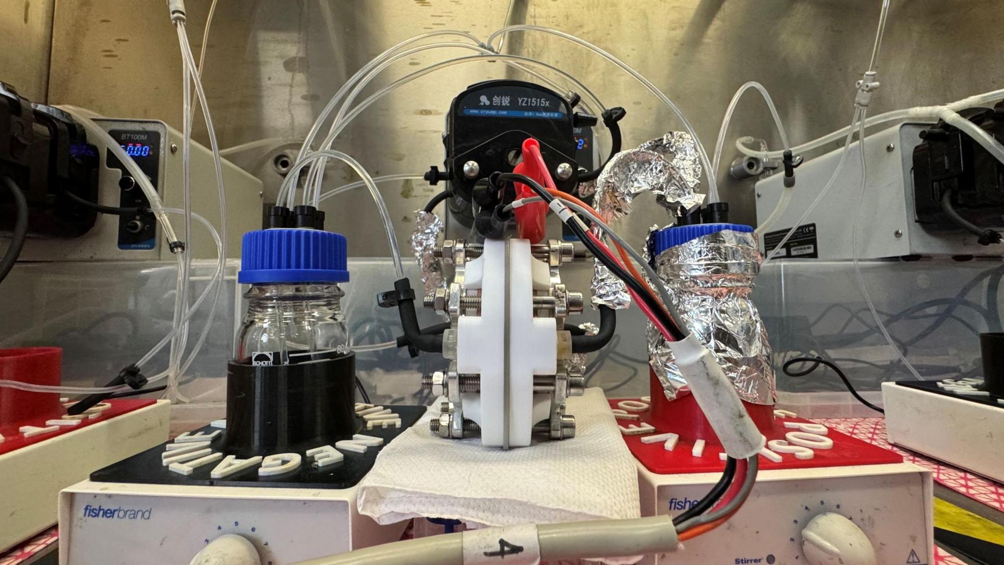 The flow battery created by Hugh O'Connor. Two clear jars with blue plastic lids are sitting on two separate platforms, one marked positive, the other negative/ The jars have tubes coming out of the top of them which are attached to a central piece of machinery that is also attached to black and red electrical wires.