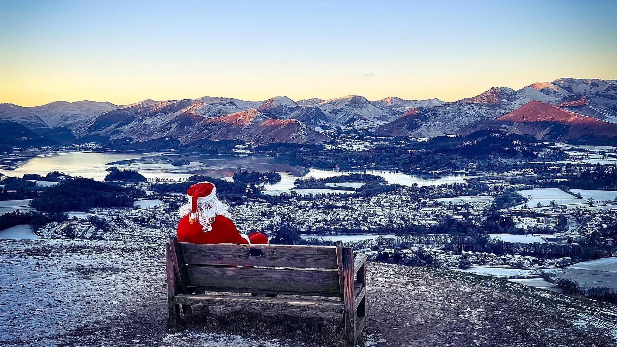 A person dressed as santa is sitting on a bench with the hills of the Lake District spread out in front of him