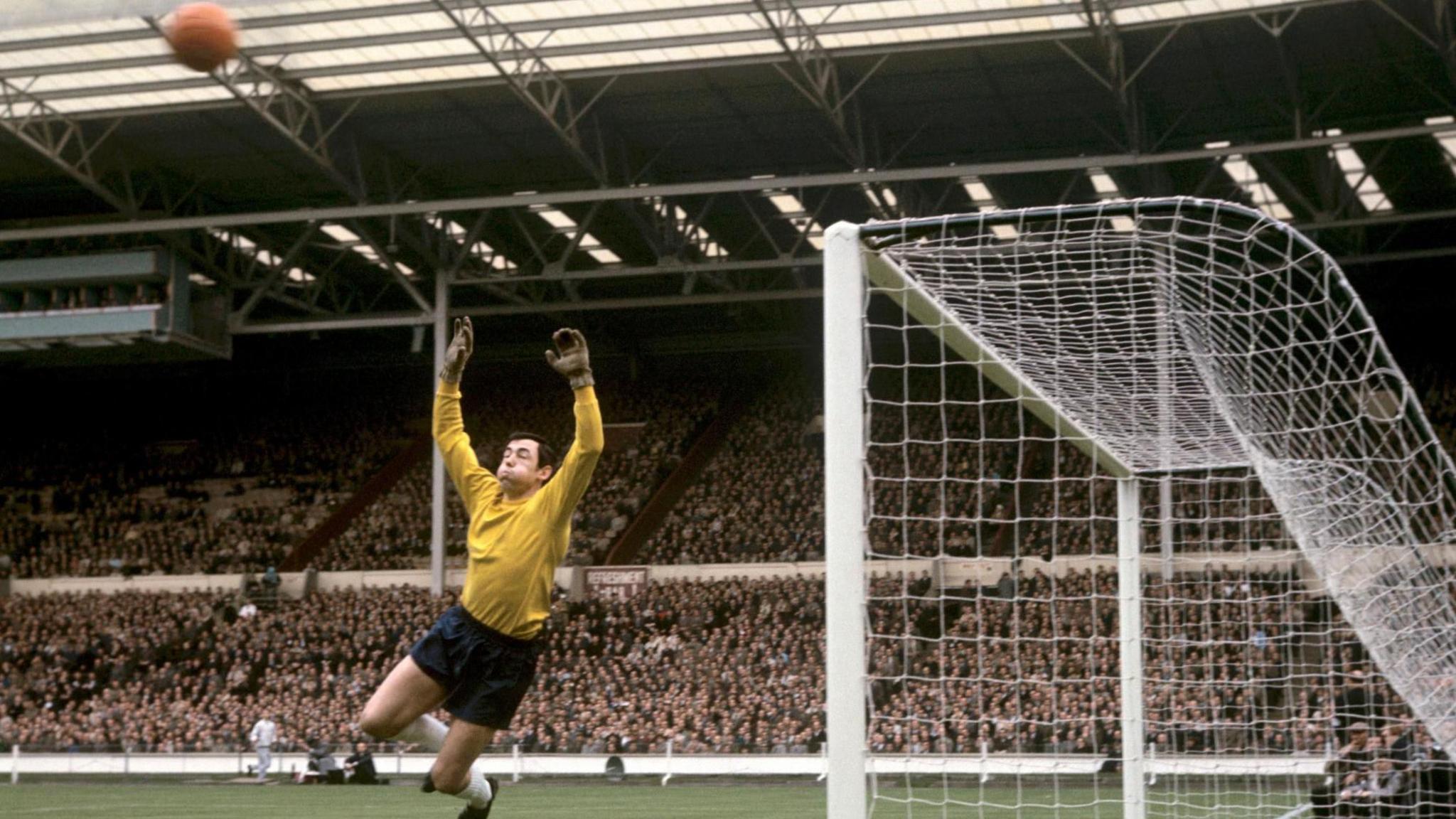 A man in a yellow jersey leaps in the air in-front of a football net in a crowded stadium