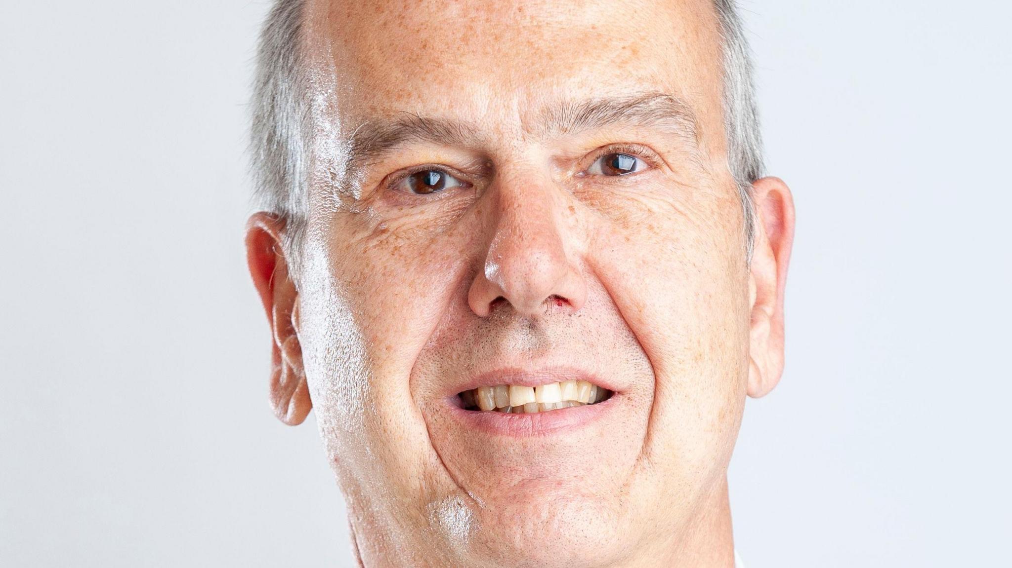 A photo showing the face of a white man with grey hair. He is stood in front of a white background
