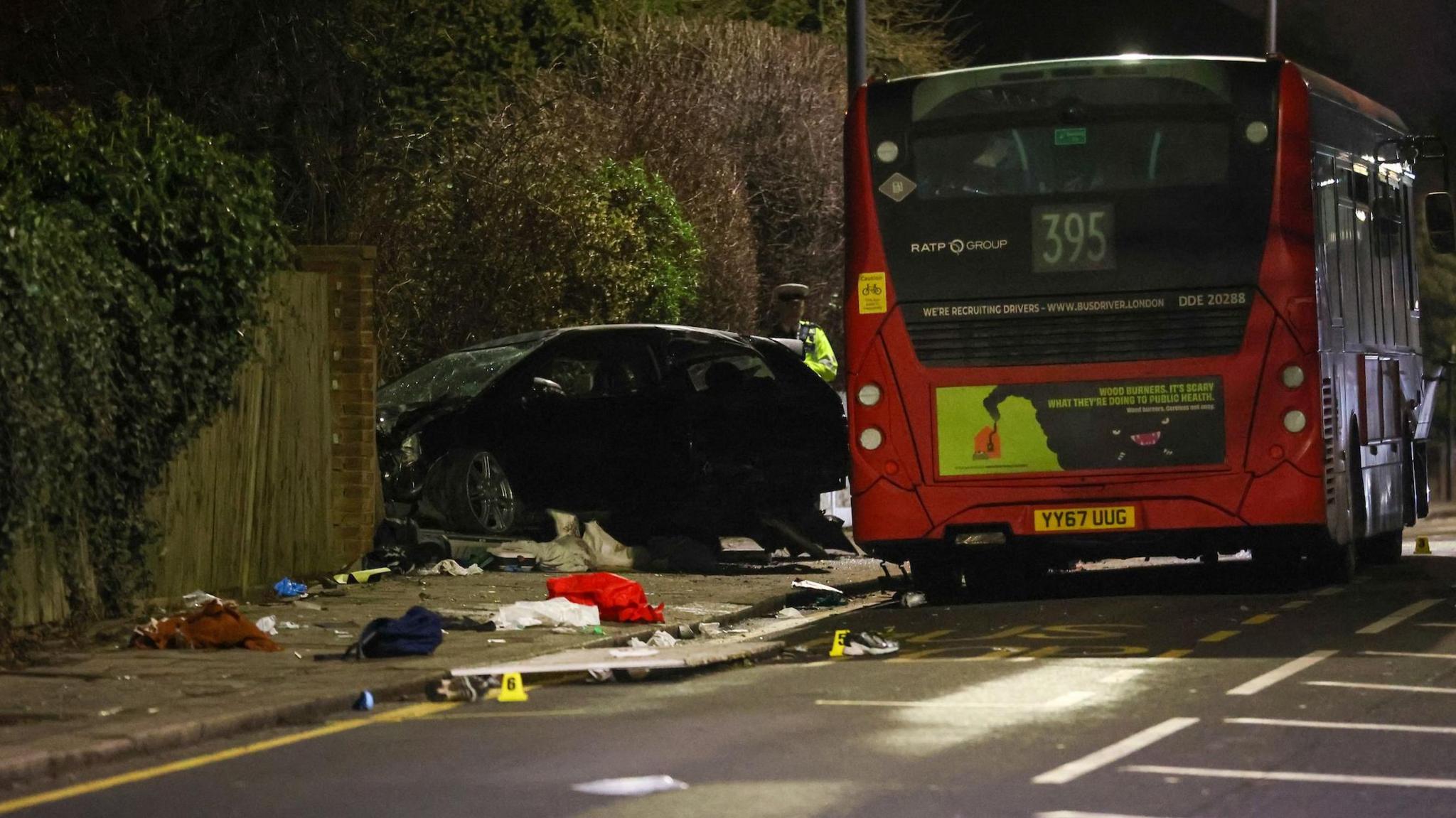 A rear view of the aftermath of the crash. The back of the bus is in view, a police officer stands near the car and litter or debris can be seen across the path.