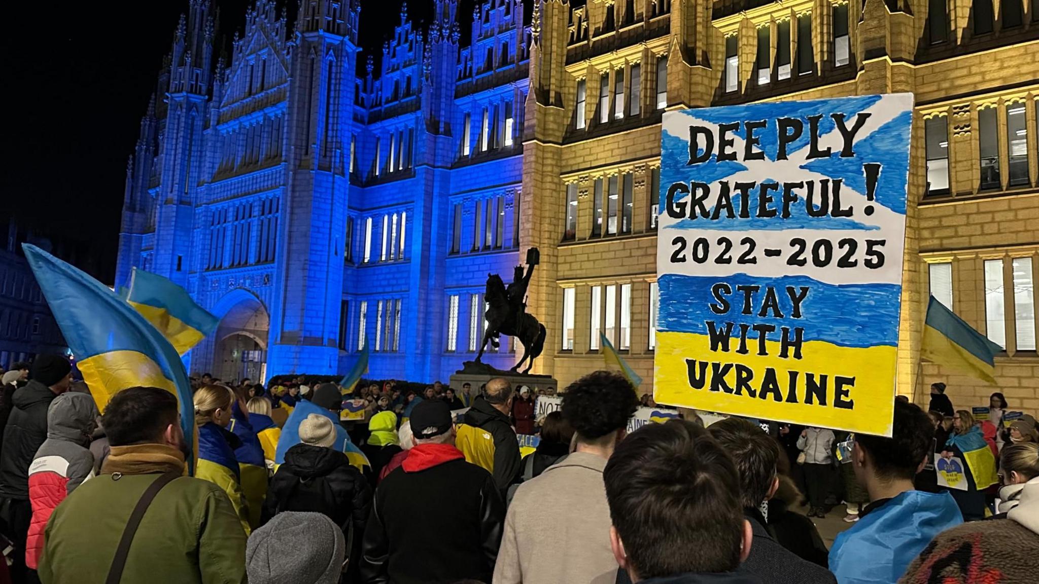 Protest in Marischal Square, Aberdeen. A poster featuring the Saltire and the Ukrainian flag reads Deeply Grateful! 2022-2025 Stay With Ukraine. Two adjacent buildings are lit in the Ukrainian colours of yellow and blue.