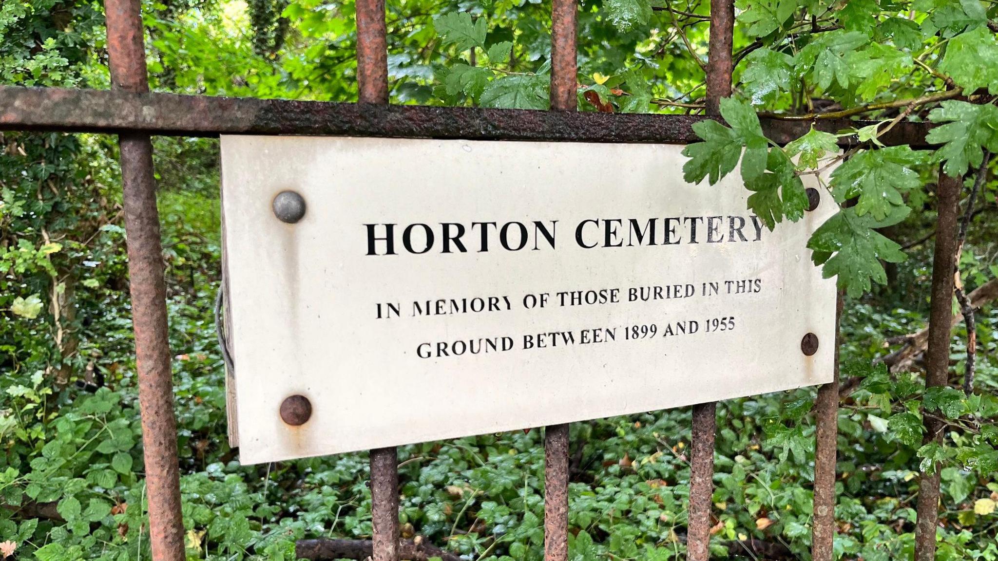 A small pale plaque attached to an iron fence. The plaque reads "Horton Cemetery, in memory of those buried in this ground between 1899 and 1955".