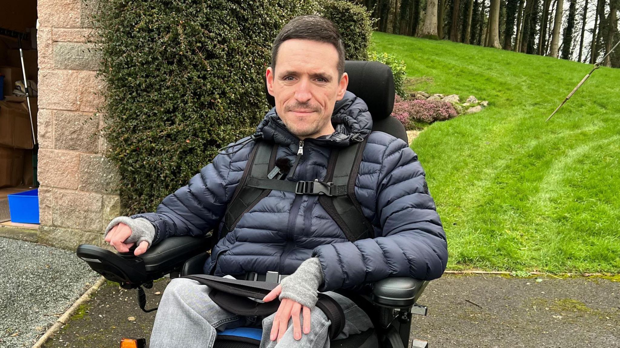 Josh Wintersgill wearing a dark blue puffer rain coat and grey jeans. He is looking at the camera beside a stone gateway entrance and a patch of grass