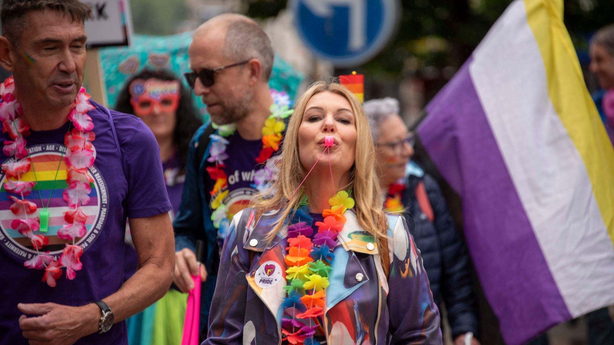 A woman wearing a purple jacket and rainbow flowers around her neck, looks at the camera while blowing a pink whistle. Other people, most wearing purple and rainbow colours, are standing around her. A purple, white and yellow striped flag is also behind her. 