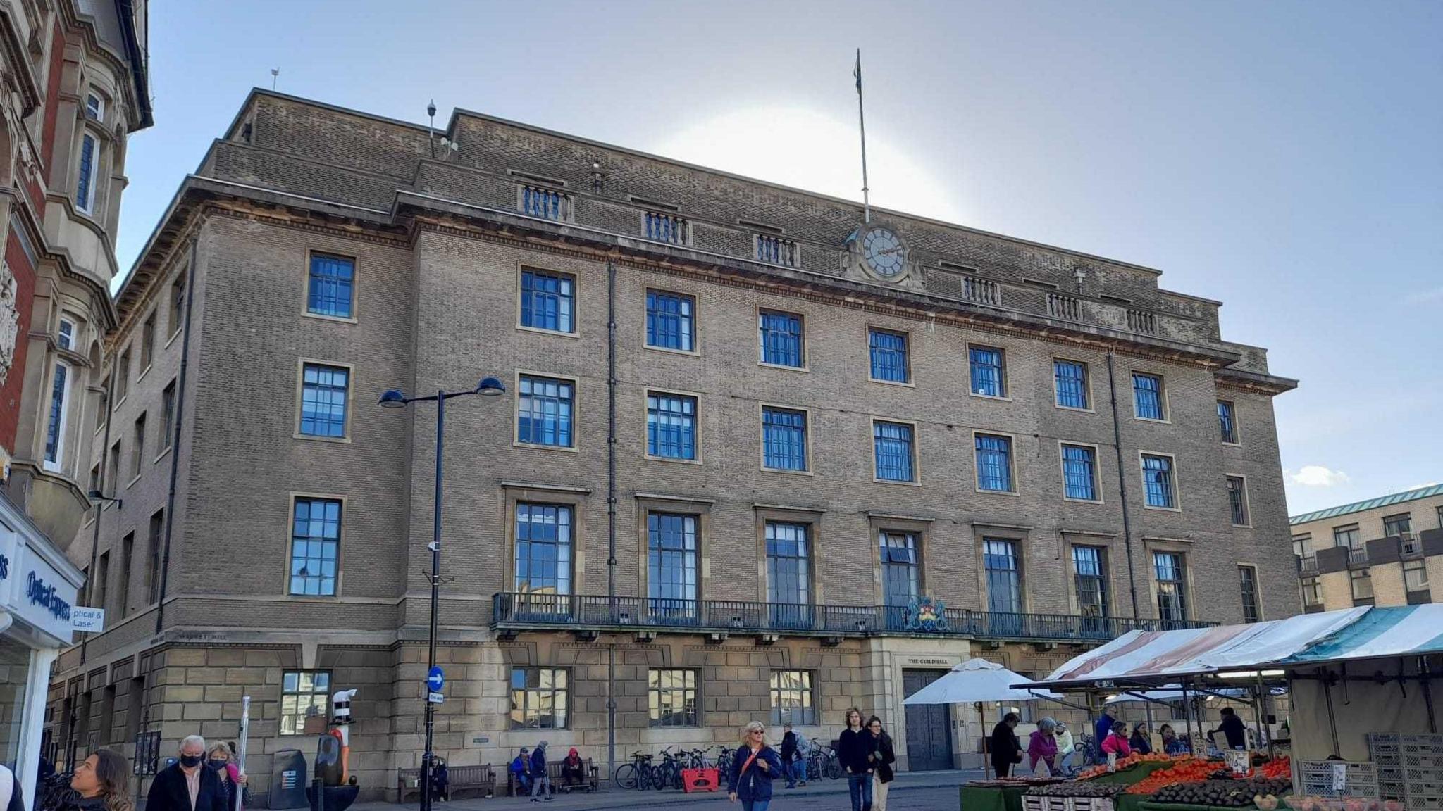 The exterior of Cambridge's Guildhall, with shops to the left and market traders directly in front of the building