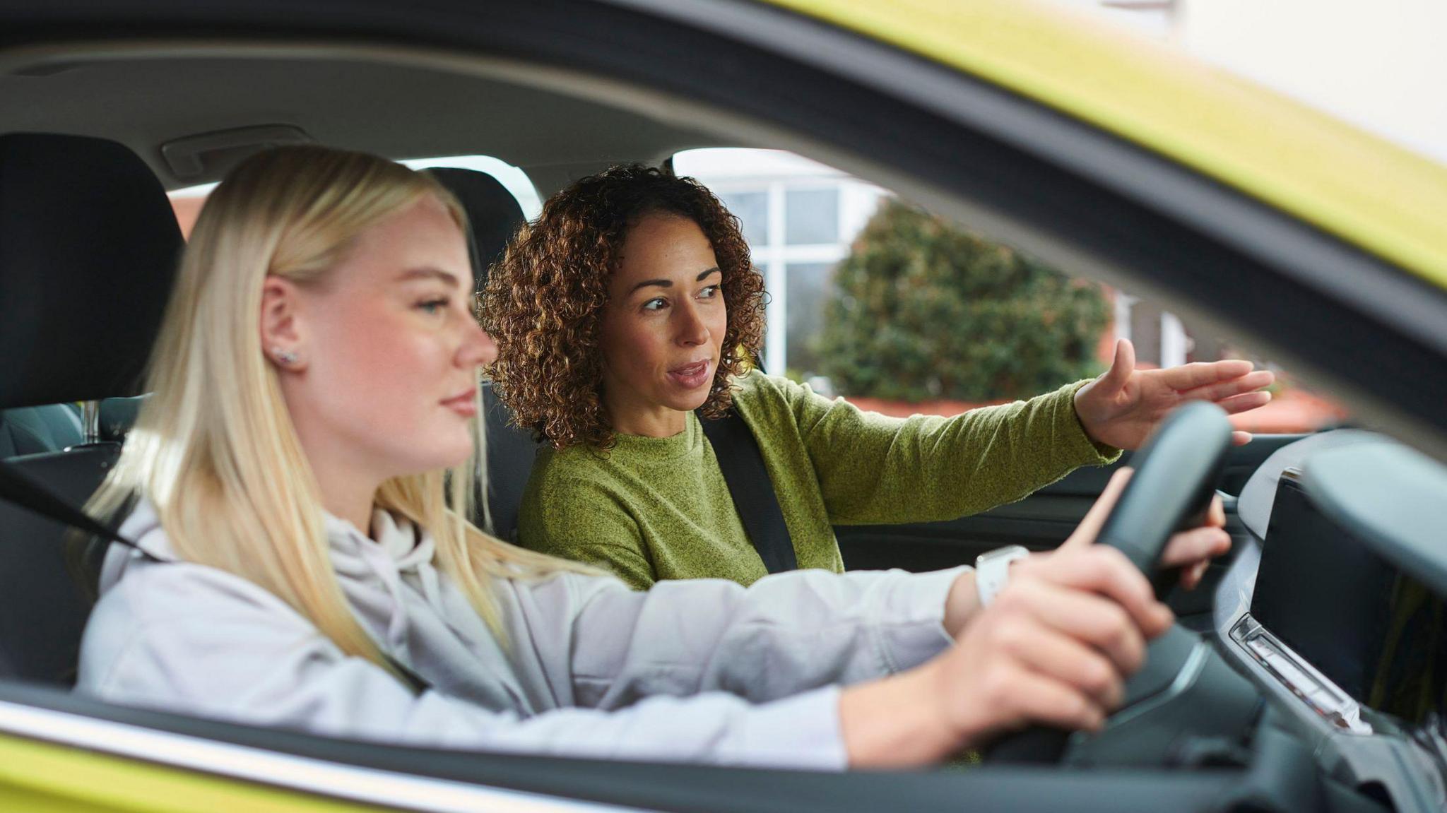 Two women in a car. The younger one is at the wheel. She is white and has blonde hair and is wearing a hoodie. An older mixed-race woman is instructing her. The car is yellow-green