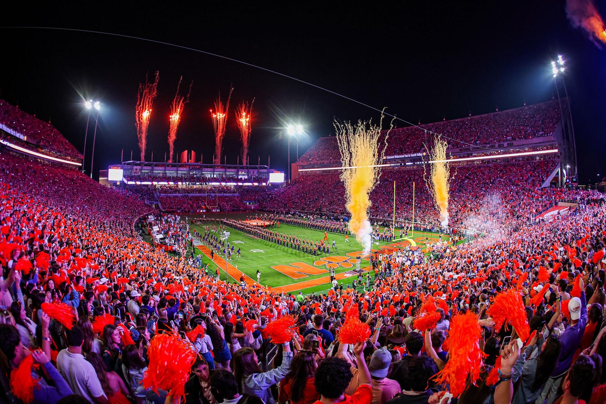 Packed football stadium at night with fans waving orange pom-poms, bright fireworks and yellow smoke rising above the field, and teams lined up near the end zone under floodlights, creating a vibrant and energetic game-day atmosphere.