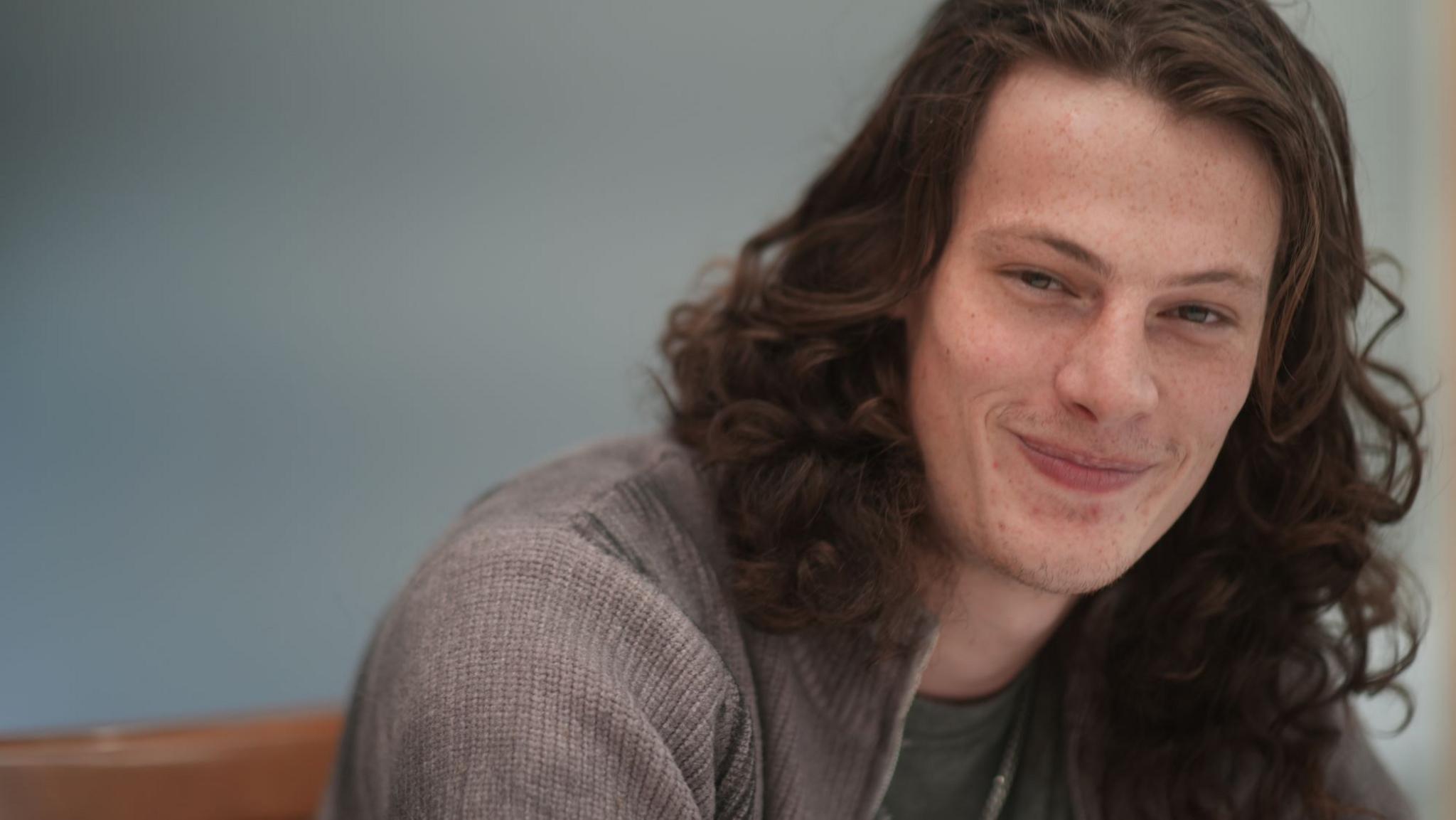 Owen Brett smiling at the camera. He has long, brown hair and is wearing a grey knitted jumper with a green T-shirt underneath. He is sat in front of a blue wall indoors.