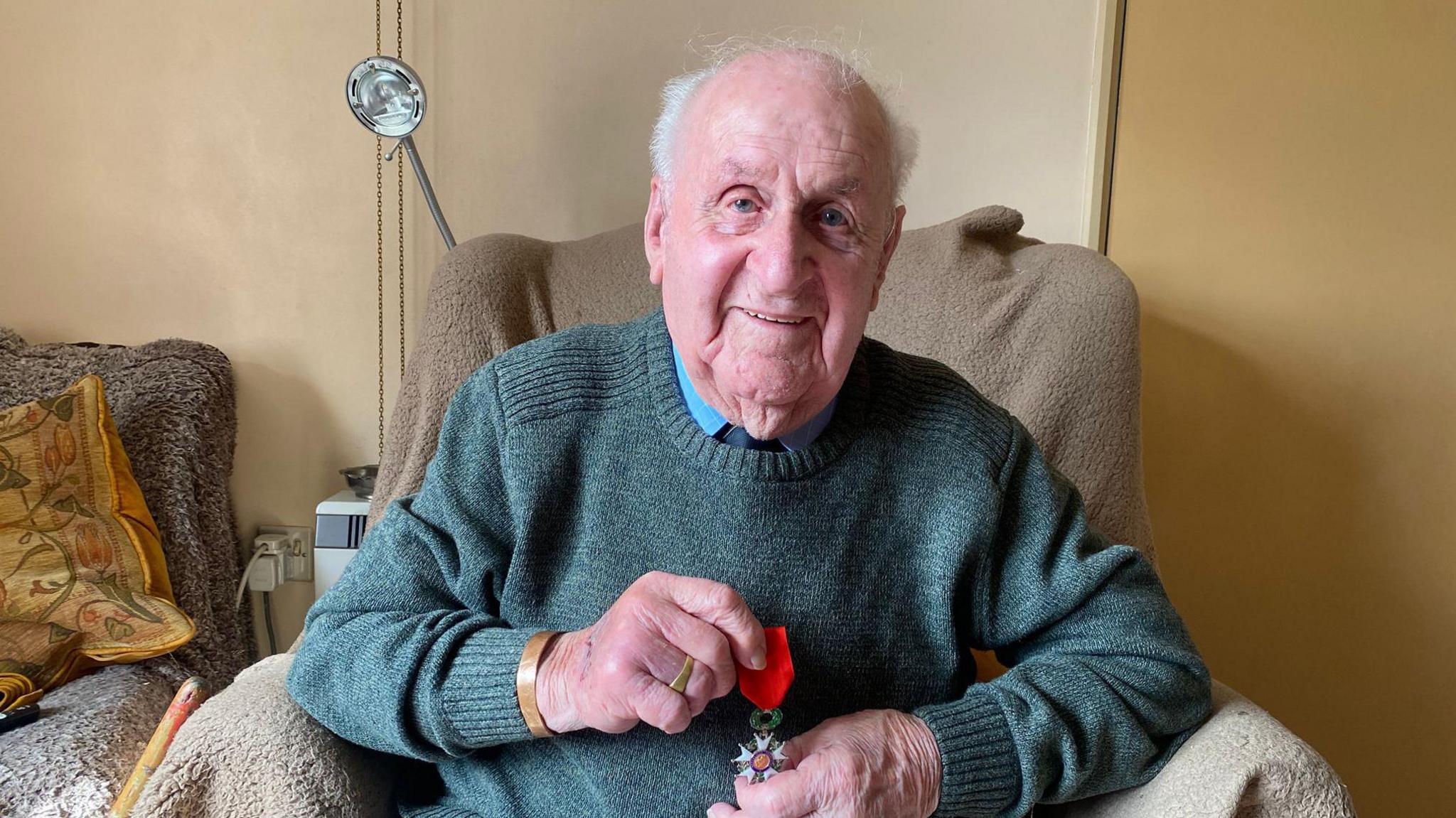 Mr Cracknell holds his Legion d'Honneur medal as his sits in a chair. He is smiling at the camera.