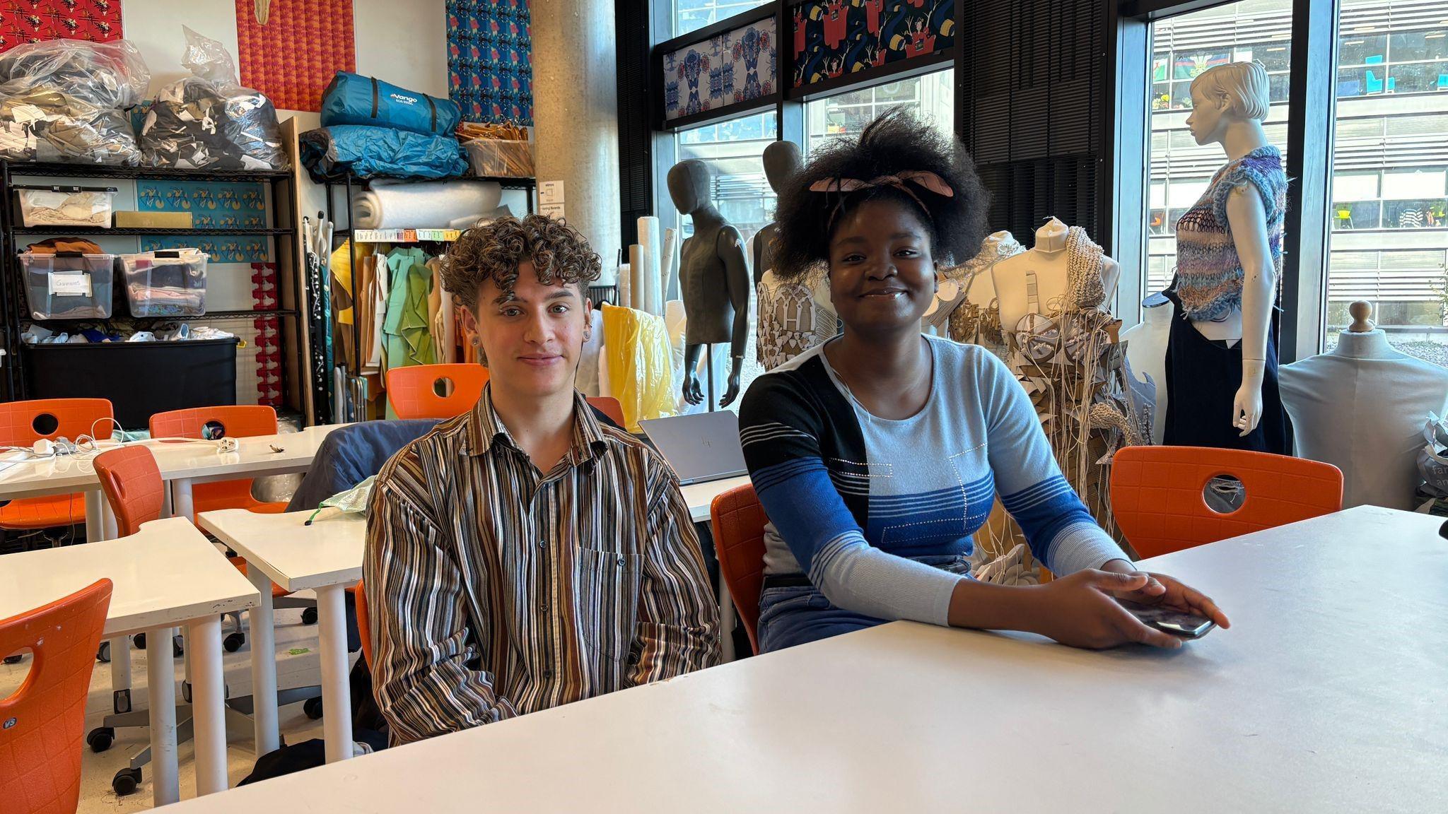 Henry Sengpiel and Debra Hart are sat at a table smiling at the camera. in the background is supplies for fashion and textile projects