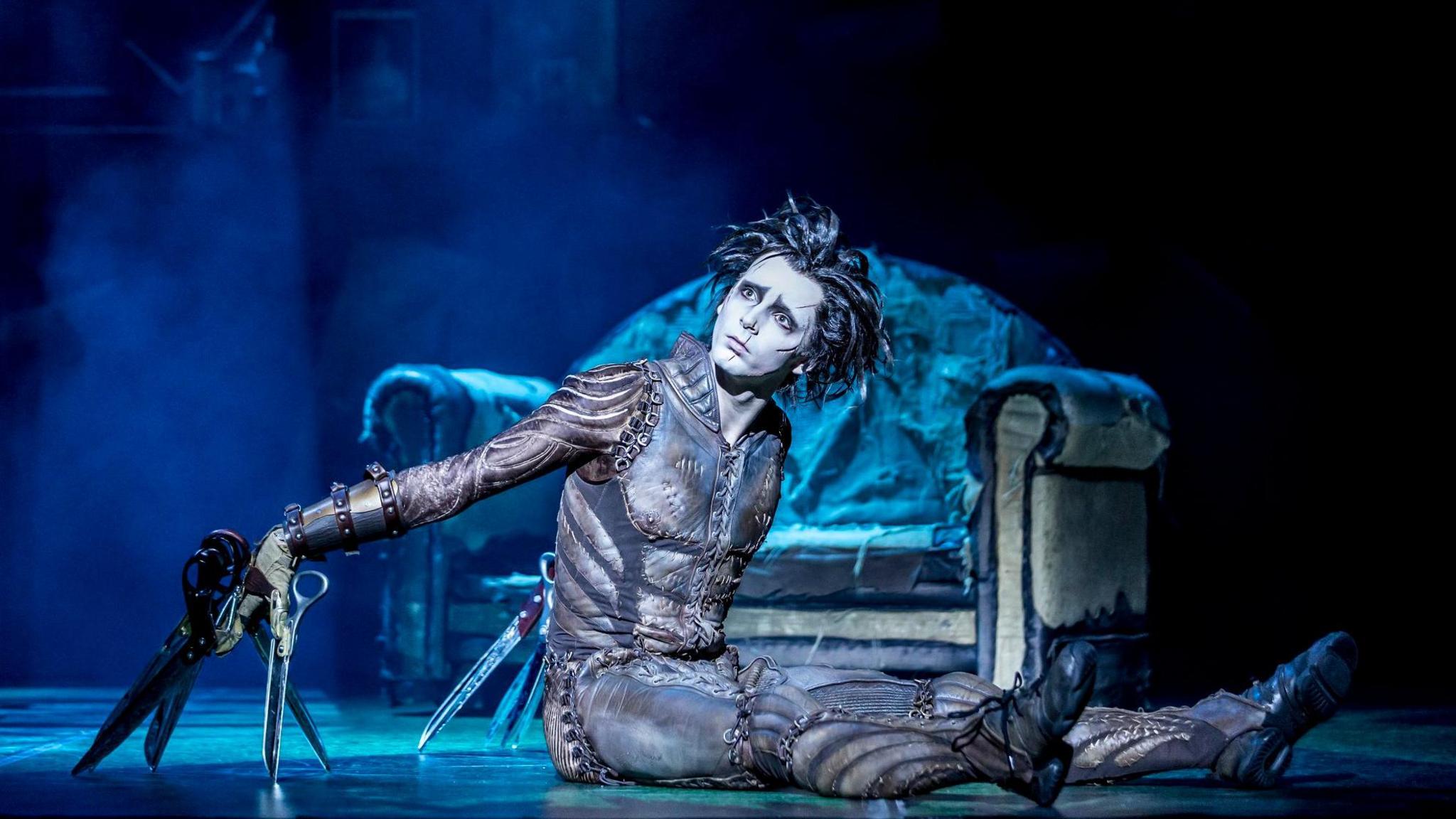 An actor playing Edward Scissorhands sits on stage at the Bristol Hippodrome in his dark costume with large pairs of scissors attached to both of his hands and a faded, tatty sofa behind him. The stage is illuminated with a blue/white light.