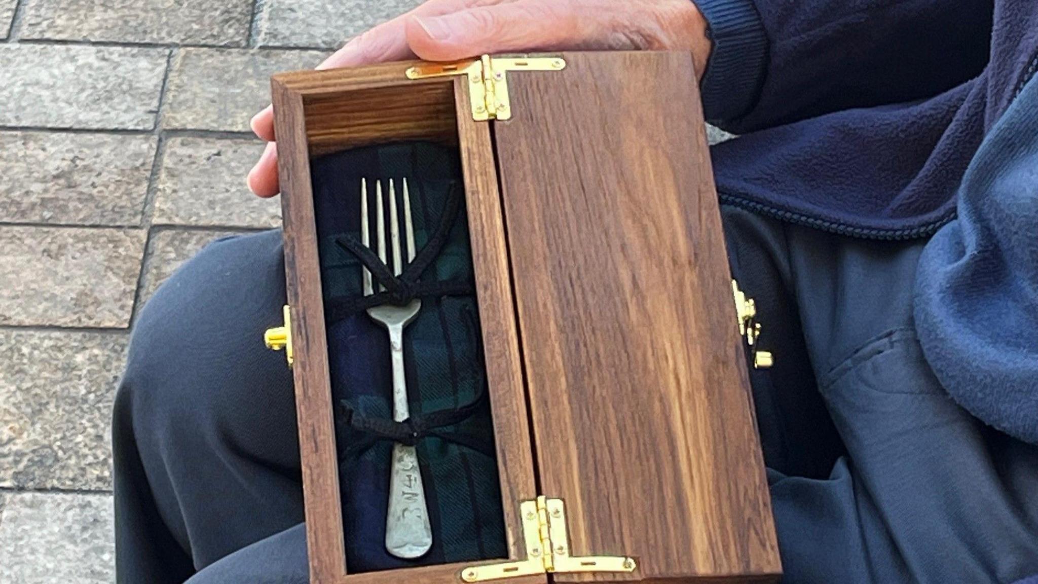 A silver-coloured fork in a brown wooden presentation box with gold-coloured hinges. It rests on David Balfour's knees.