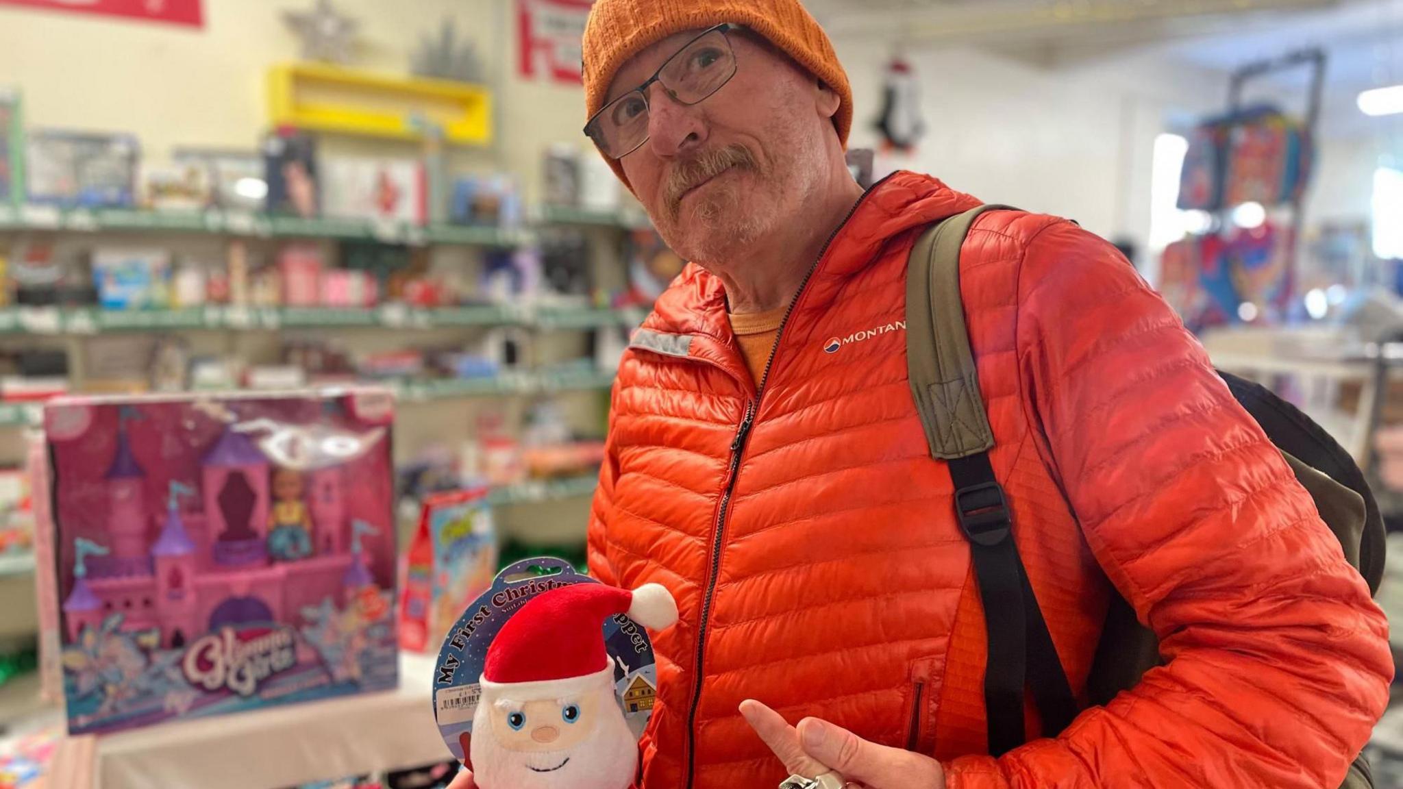 A man wearing an orange hat and orange jacket points at a small Santa.