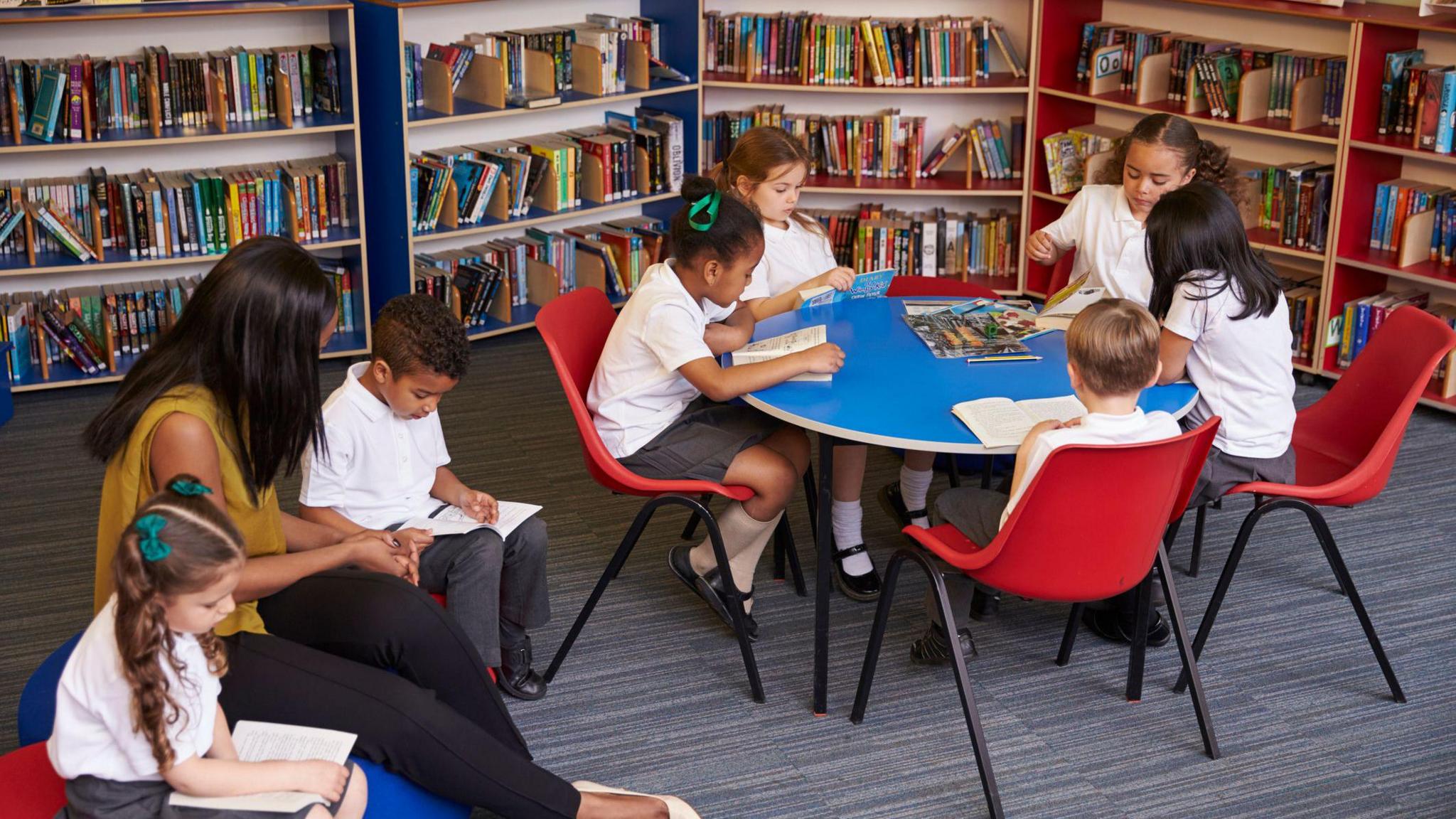 Children in white polos and a teacher sit around a table and on soft cushions reading books in a library