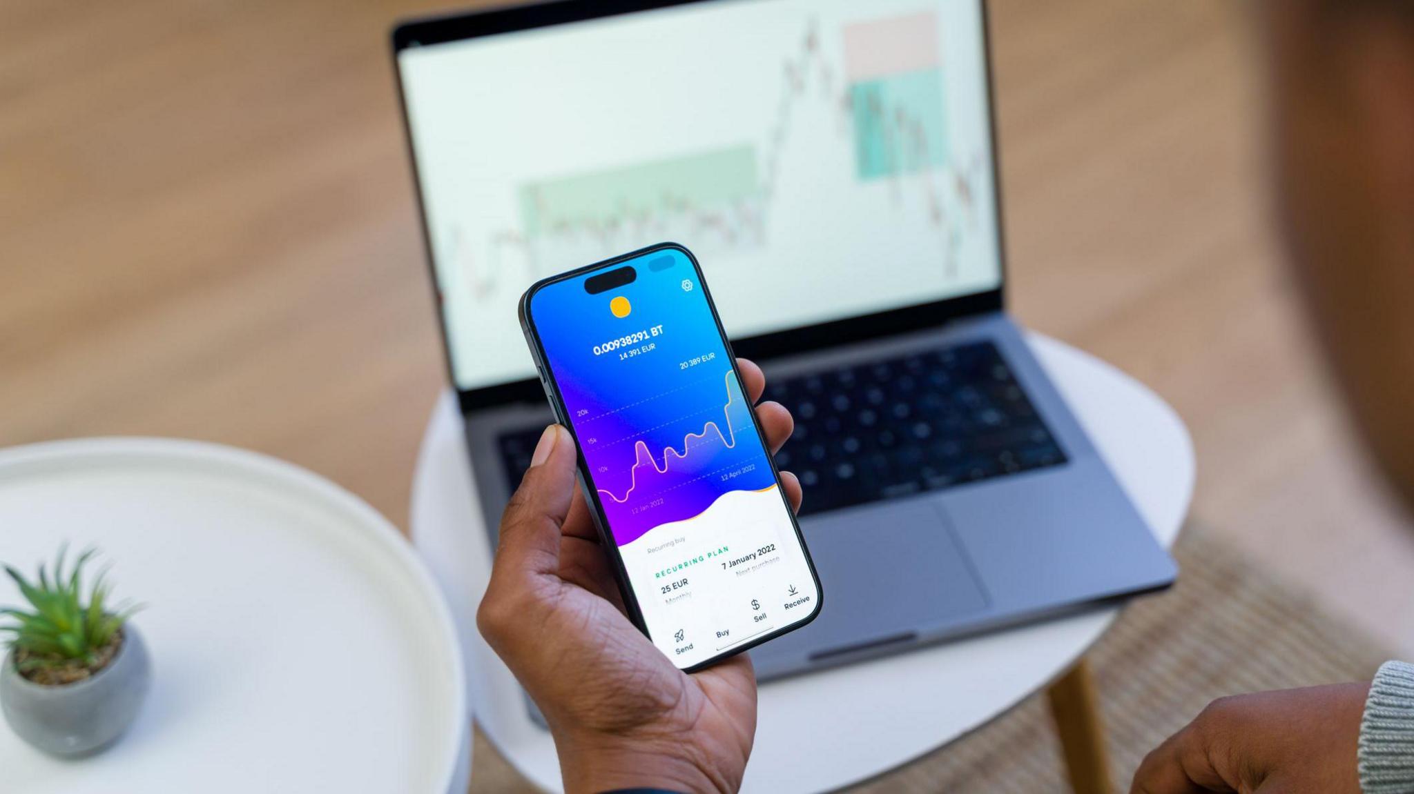 Trader's hand holding a phone showing financial information with a laptop on a similar page behind.
