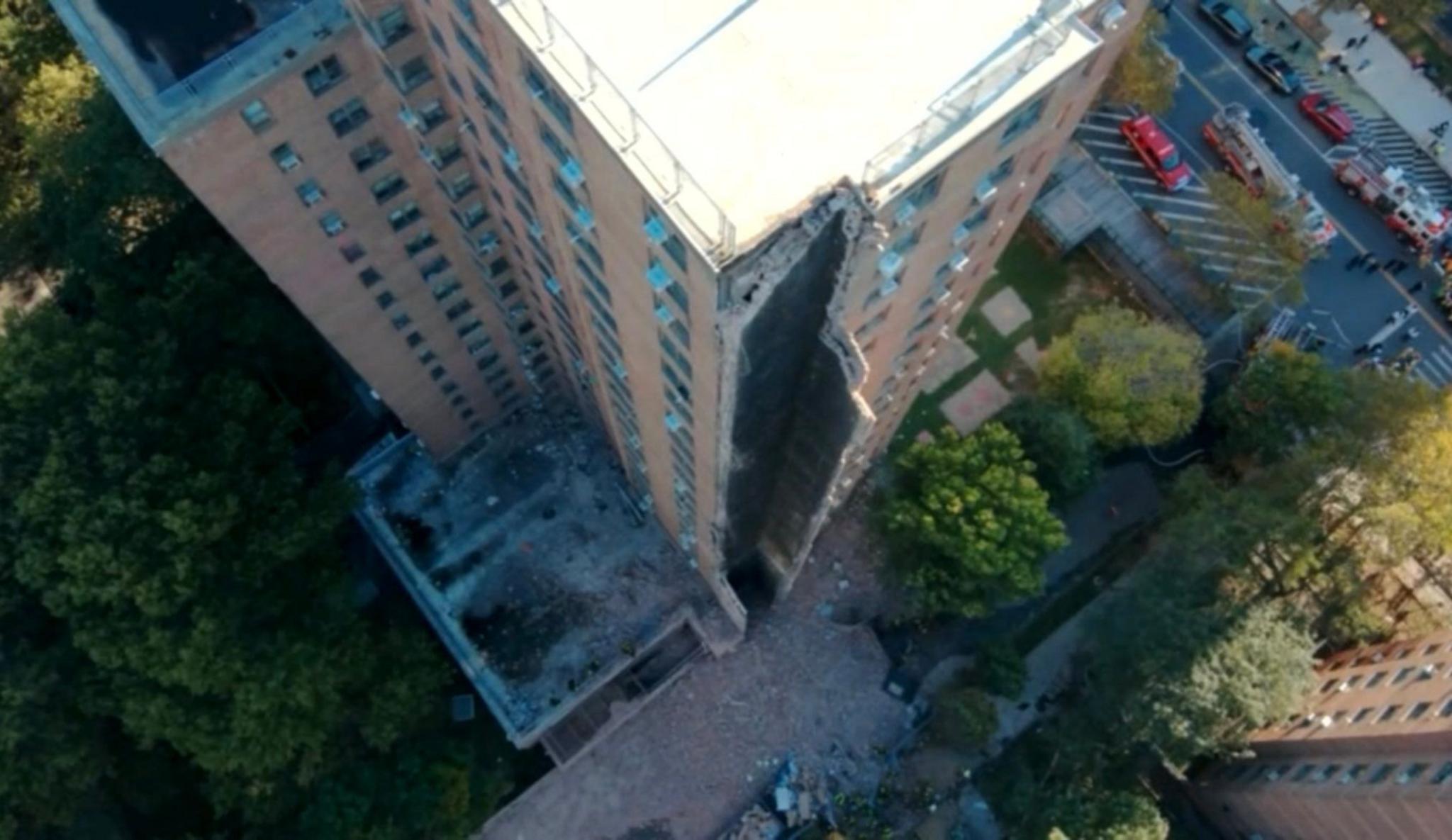 Aerial pic of a large brick apartment building with a section in the middle of the frame missing. The roof is white and trees on the ground surround the building