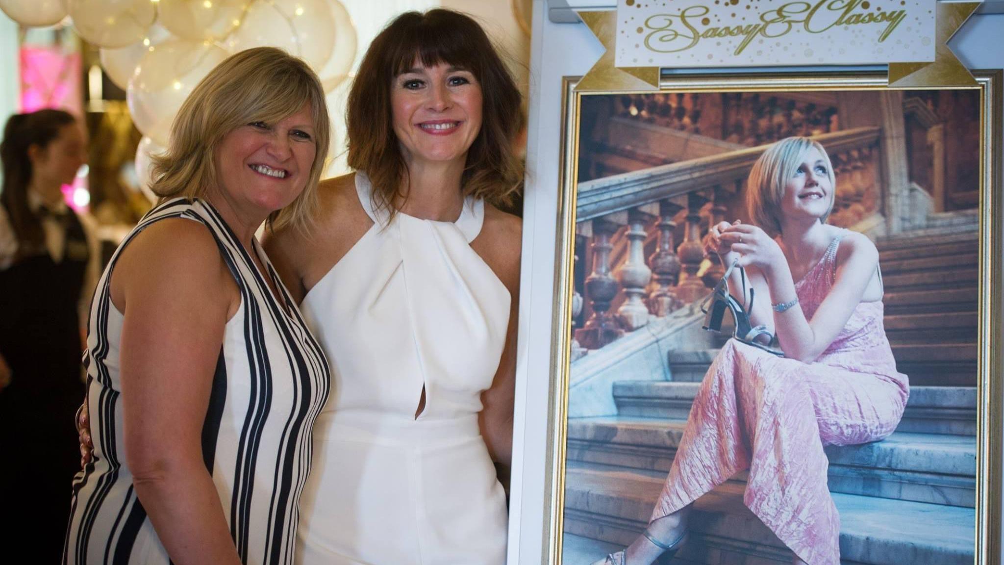 Smiling Angela MacVicar and Lindsay MacCallum wearing party dresses posing next to a photograph of Johanna MacVicar with helium-filled balloons behind them