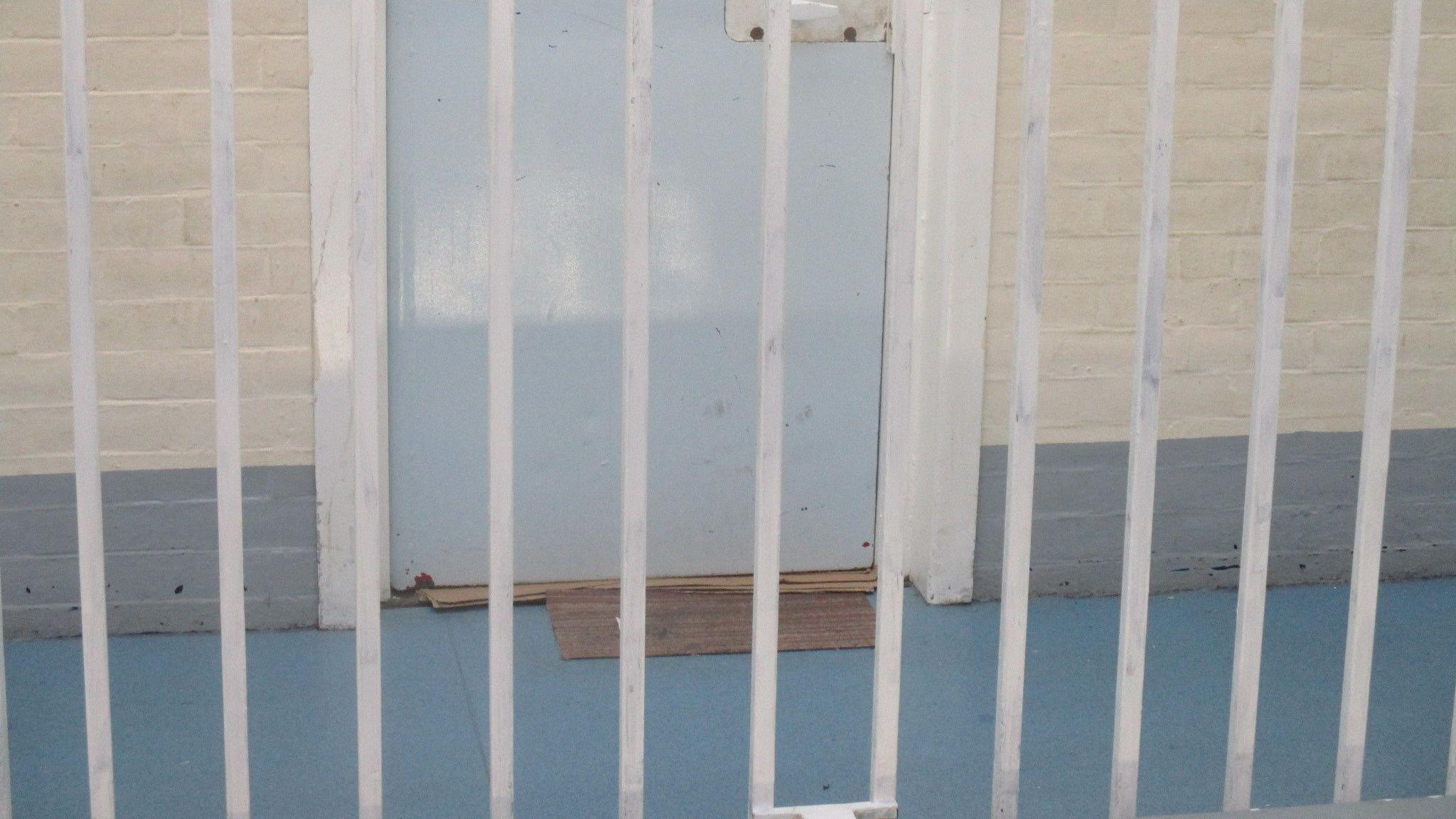 The outside of a cell door is seen from between prison bars. There are several pieces of cardboard wedged in to the gap under the door.