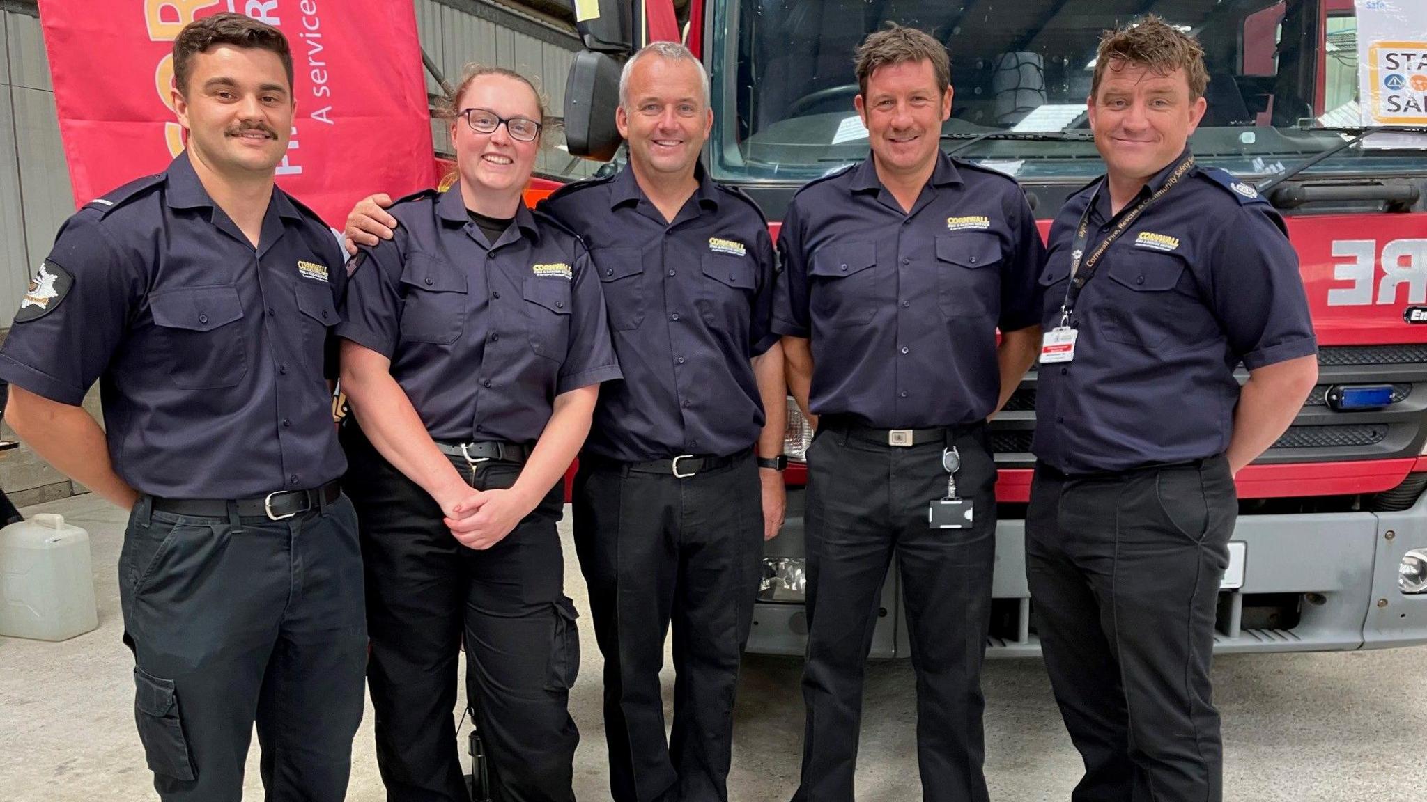 Five uniformed members of Cornwall Fire and Rescue Service. Four are men and one is Ms Thomas. They are standing in front of a fire engine in the event hall.