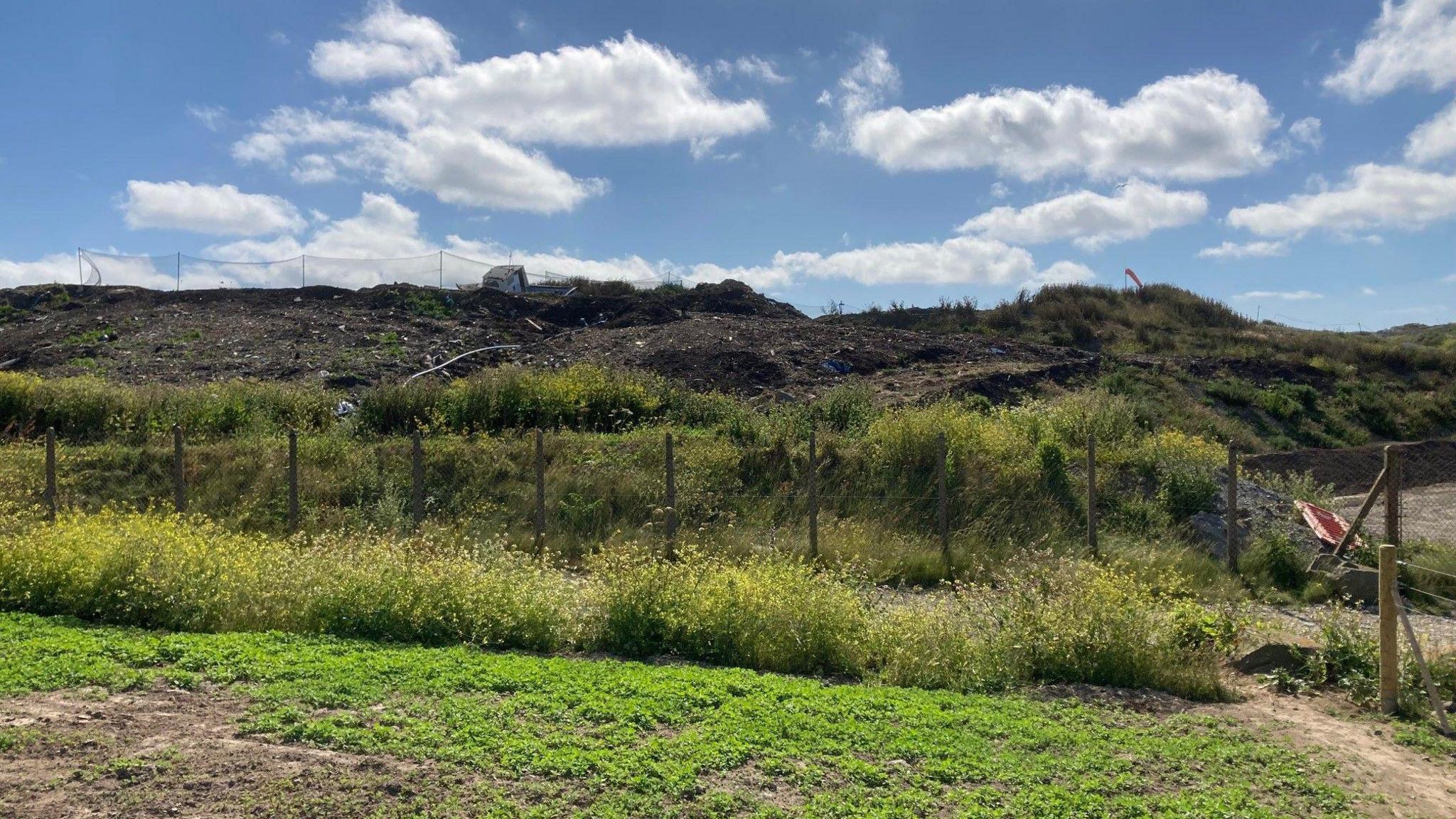 A landfill site showing a mound of dirt littered with rubbish including a boat. It is a partly cloudy day.