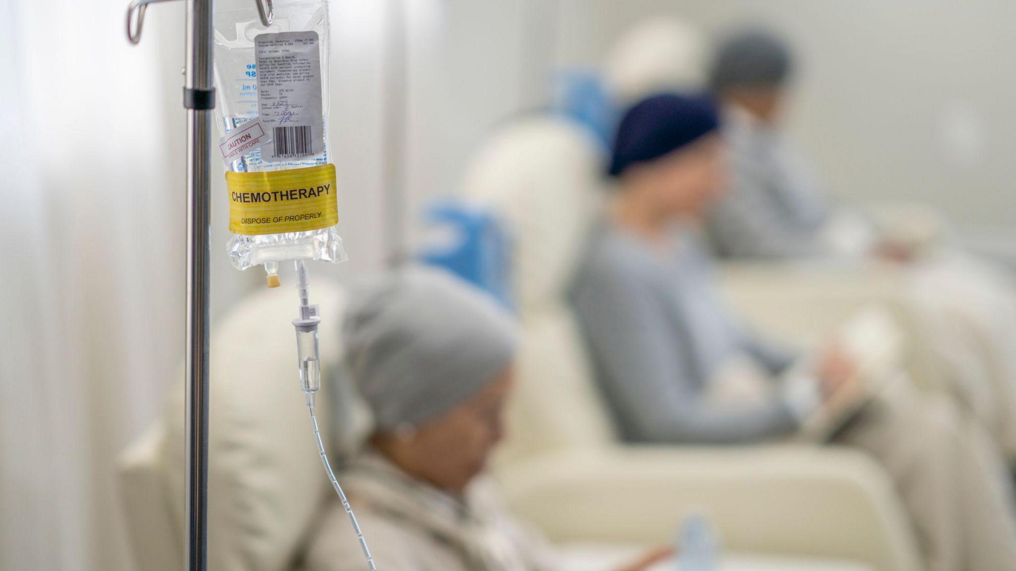 An IV bag with clear liquid in it with a yellow sticker saying "chemotherapy". Three patients can be seen sitting in chairs behind it.