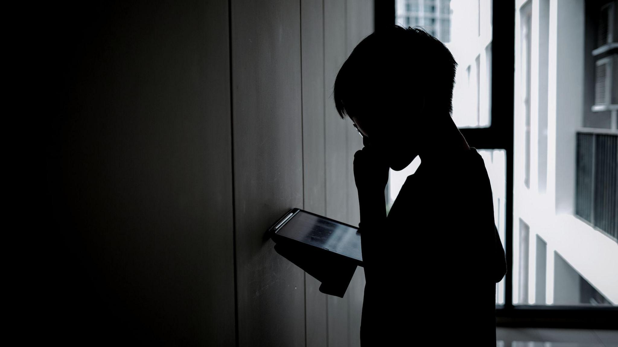 A boy stands in the corner of a room playing with a computer tablet. It is dark.