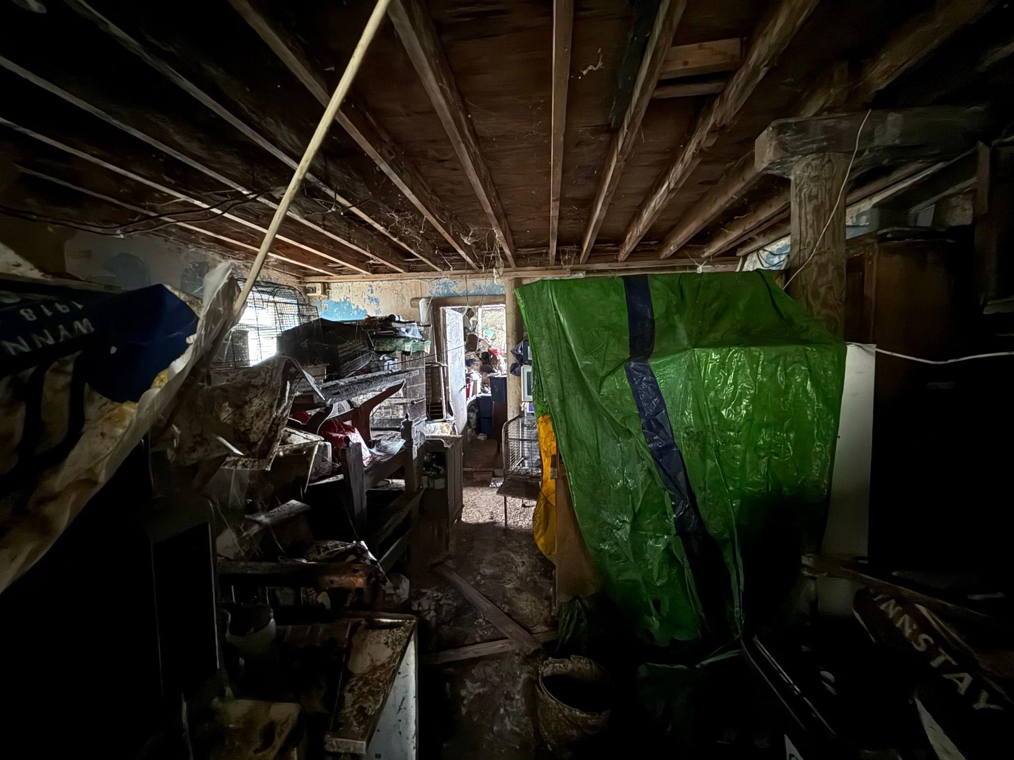 A dark image of lots of clutter in what looks like a living room. On the left, there is a large green sheet which seems to be covering a pile of clutter. The room looks very dirty. 